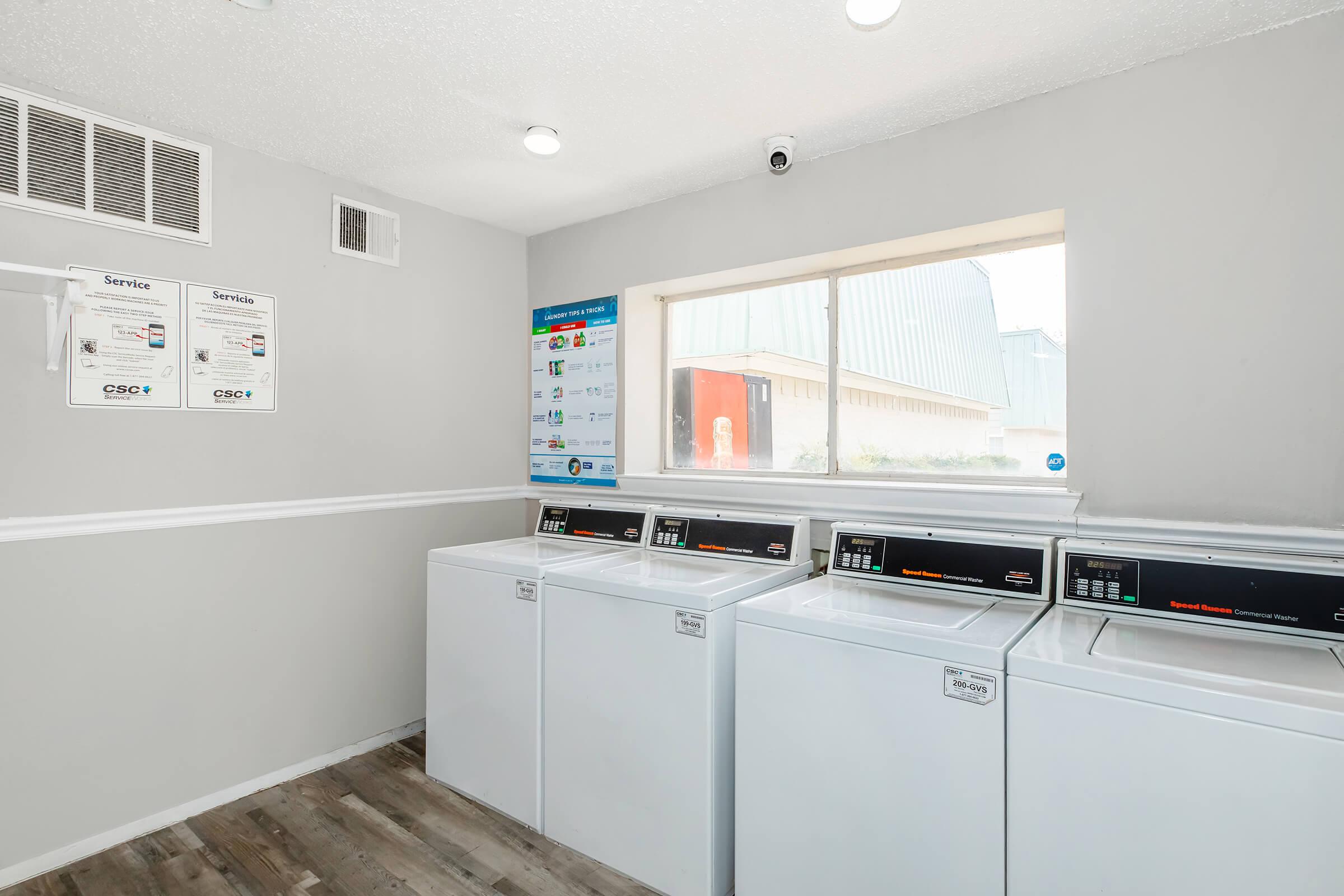 A clean and well-lit laundry room featuring three white washing machines lined up against a gray wall. There are service information posters on the wall, and a window providing natural light, with a view of the outside.