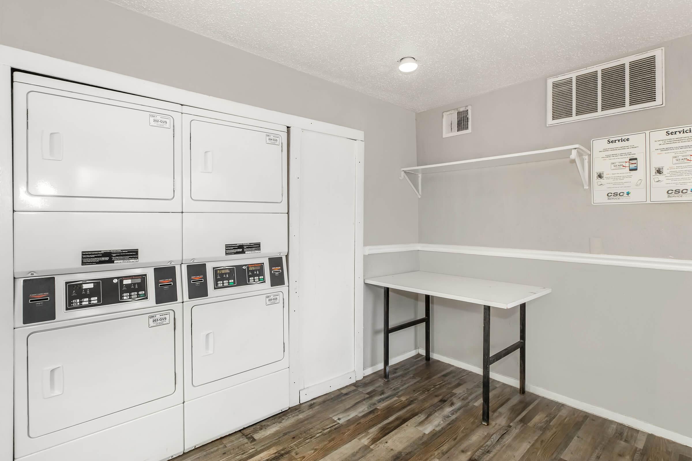 A clean and modern laundry room featuring stacked coin-operated washing machines and dryers on the left, with a table for folding clothes. The walls are painted light grey, and there is a shelf with service information displayed above the table.