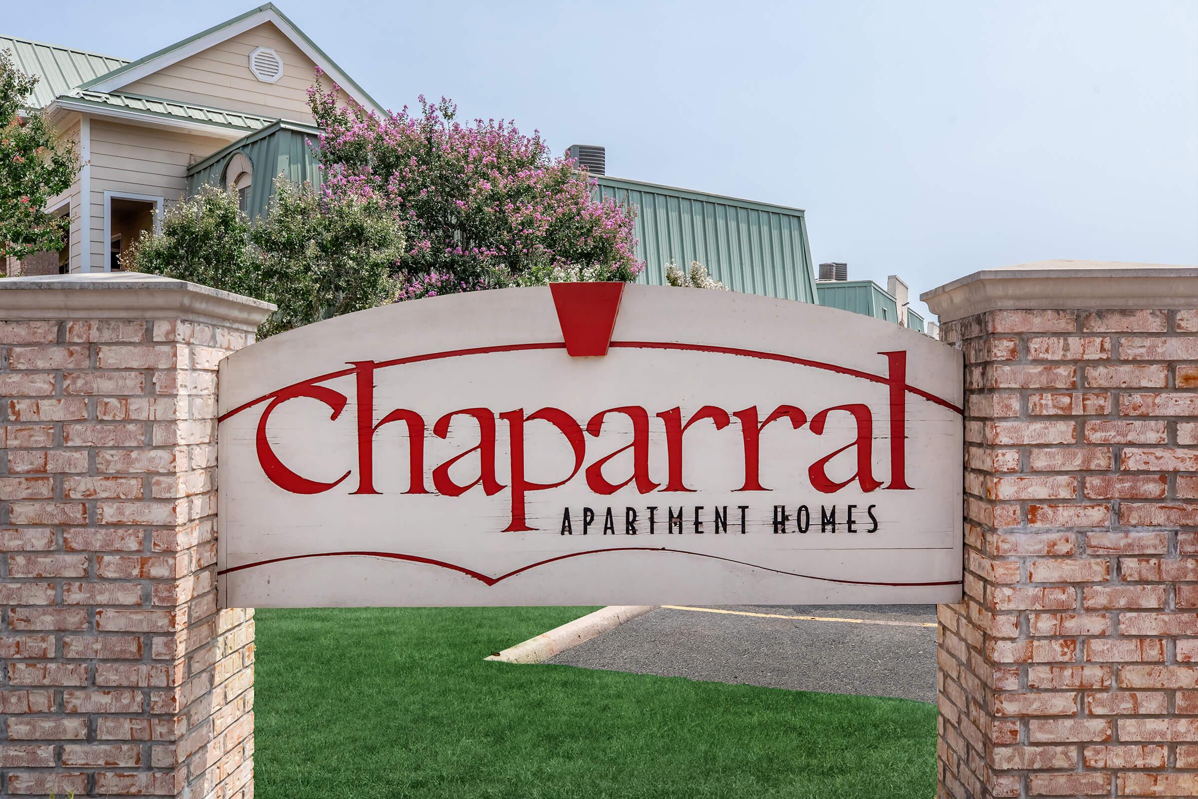 Sign for Chaparral Apartment Homes, featuring a large red logo with the name "Chaparral" prominently displayed. The sign is set against a backdrop of green grass and a blurred building in the background.