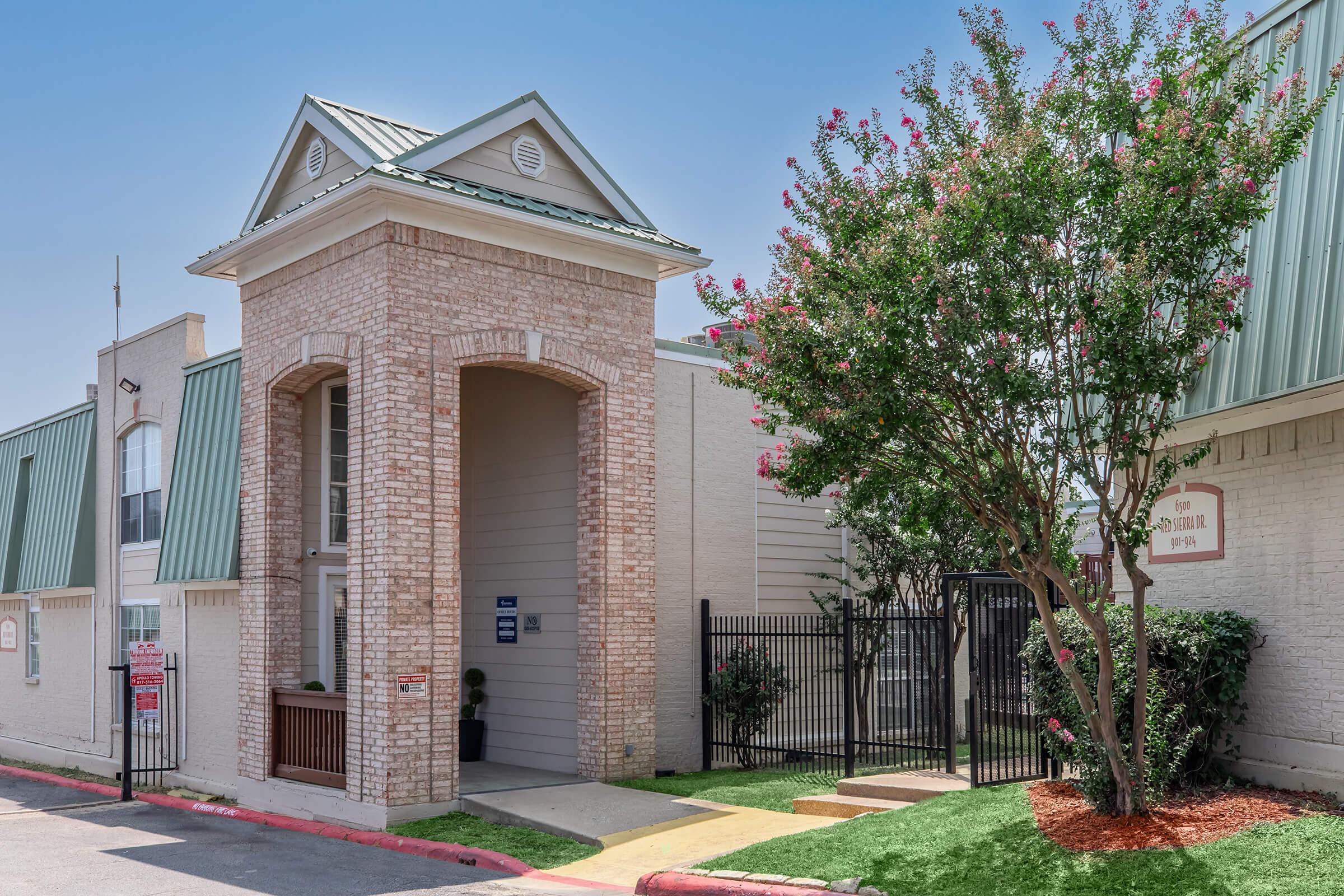 A brick building with a peaked entrance, featuring a small sign, surrounded by greenery and a fence. The sky is clear and blue, and there are flowering bushes near the entrance. The structure has a light-colored facade with a green roof, indicating a welcoming entryway to a property or facility.