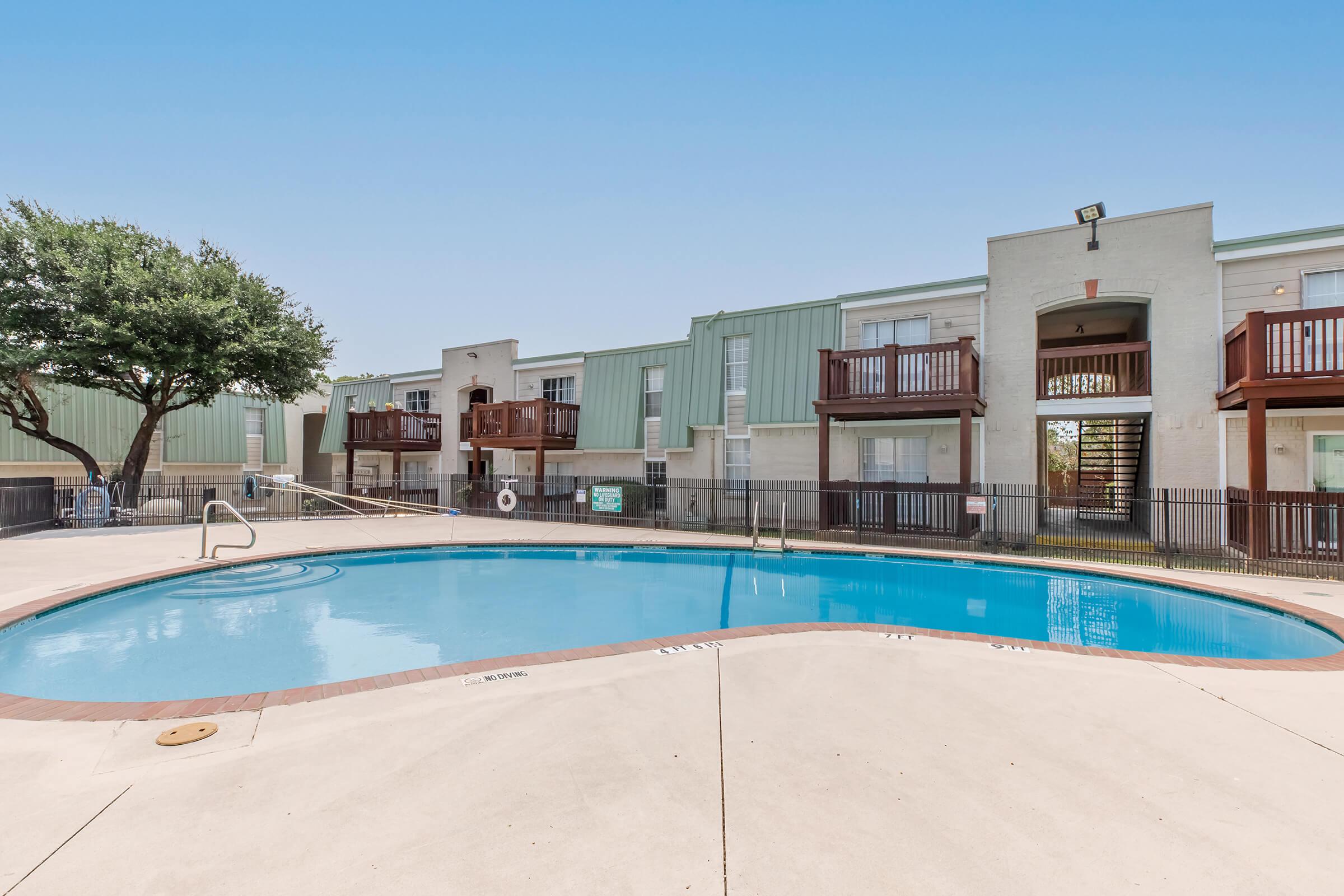 A clear view of a swimming pool surrounded by a patio area, with nearby apartment buildings featuring balconies. The scene is bright and sunny, showcasing a well-maintained community space. Lush trees are visible in the background, enhancing the relaxing atmosphere.