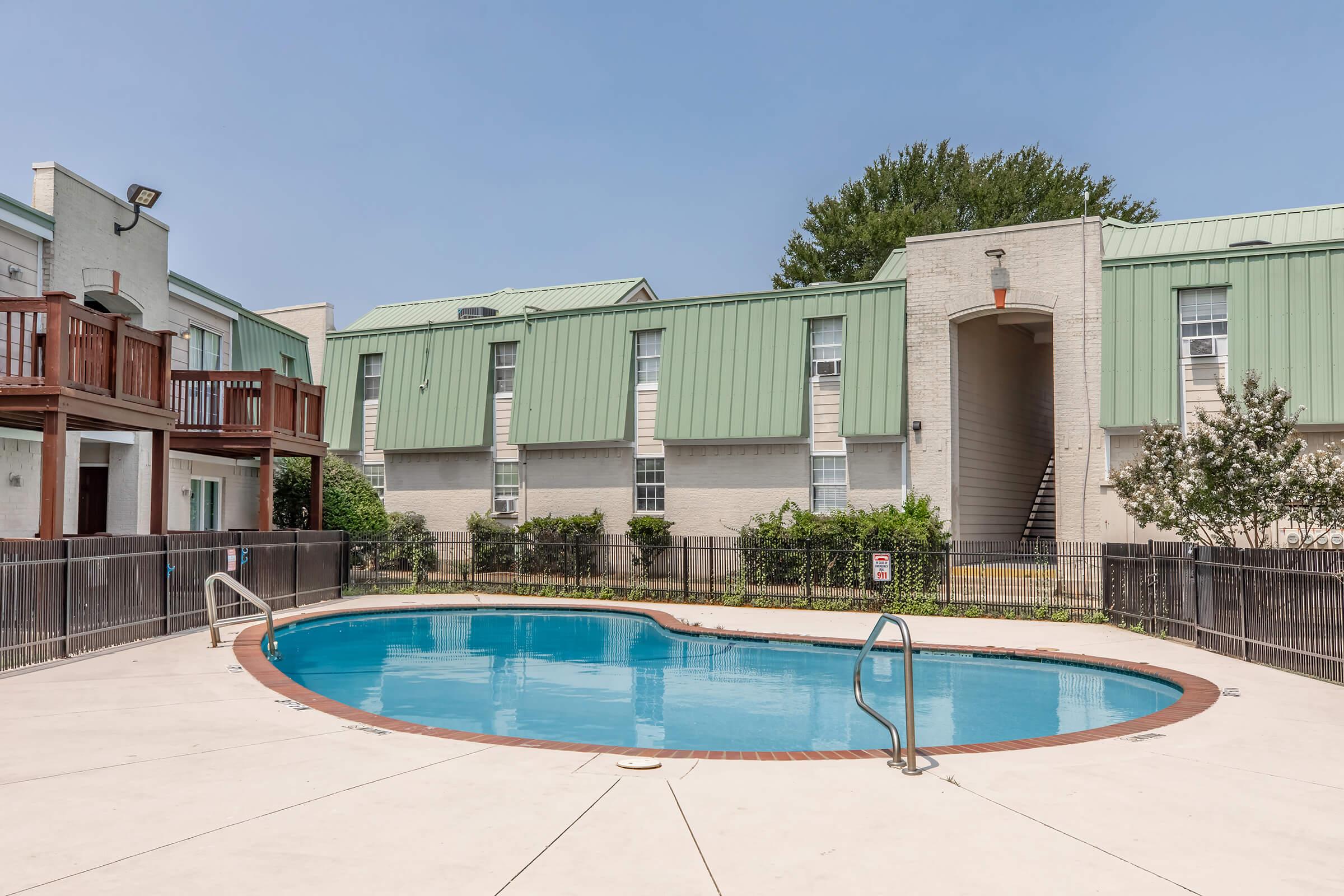 Swimming pool in a residential area with a curved shape, surrounded by a fenced patio. The area features green-roofed buildings, trees in the background, and a clear blue sky above. Stairs lead up to an upper level of the building. A sign near the pool indicates safety rules.