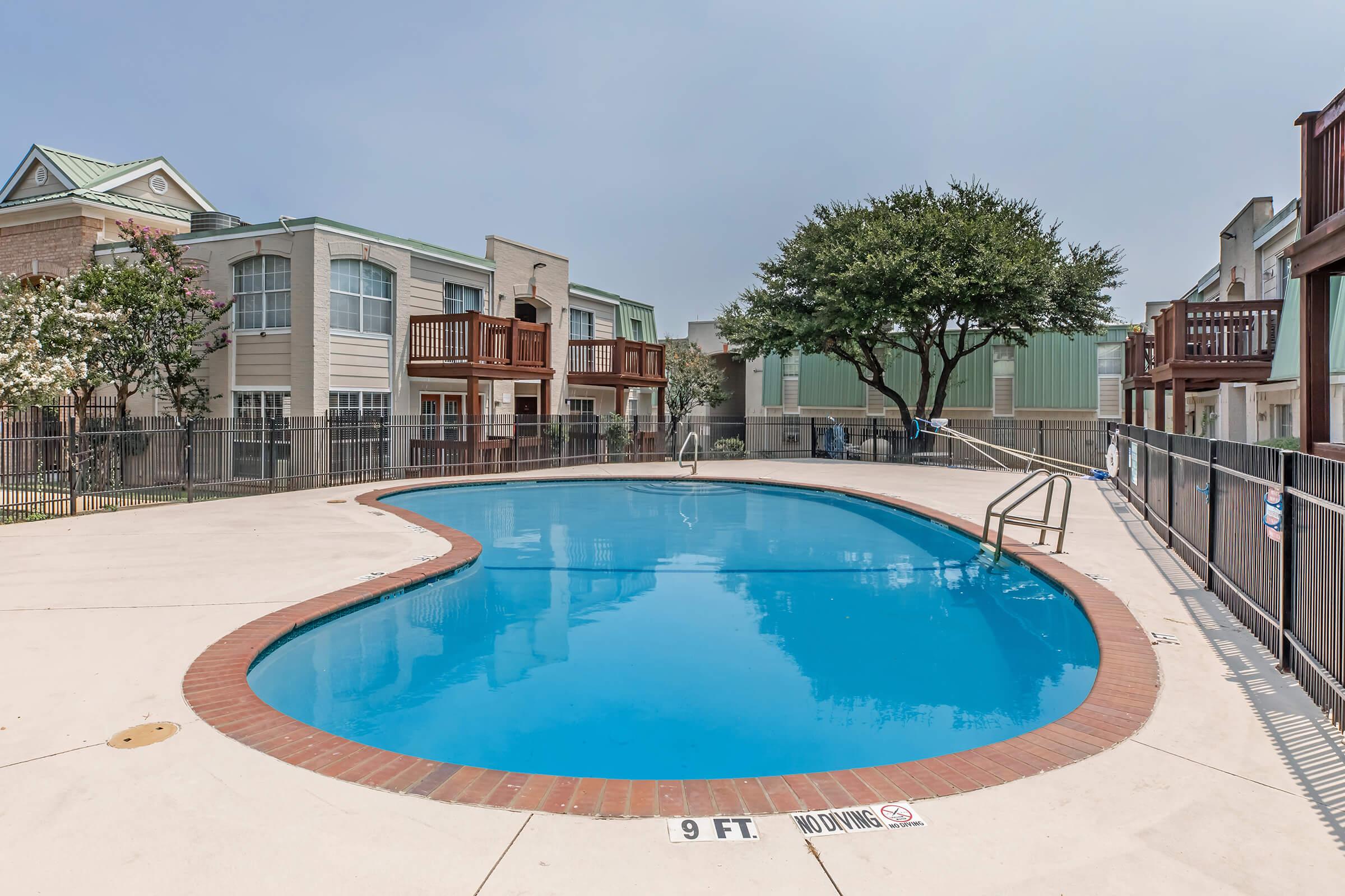 A clear, blue swimming pool in an outdoor area surrounded by a fence. Nearby are several multi-story residential buildings with balconies. The pool features a ladder and a "9 FT NO DIVING" sign on the pool deck. Lush trees provide shade around the area, creating a relaxing atmosphere.