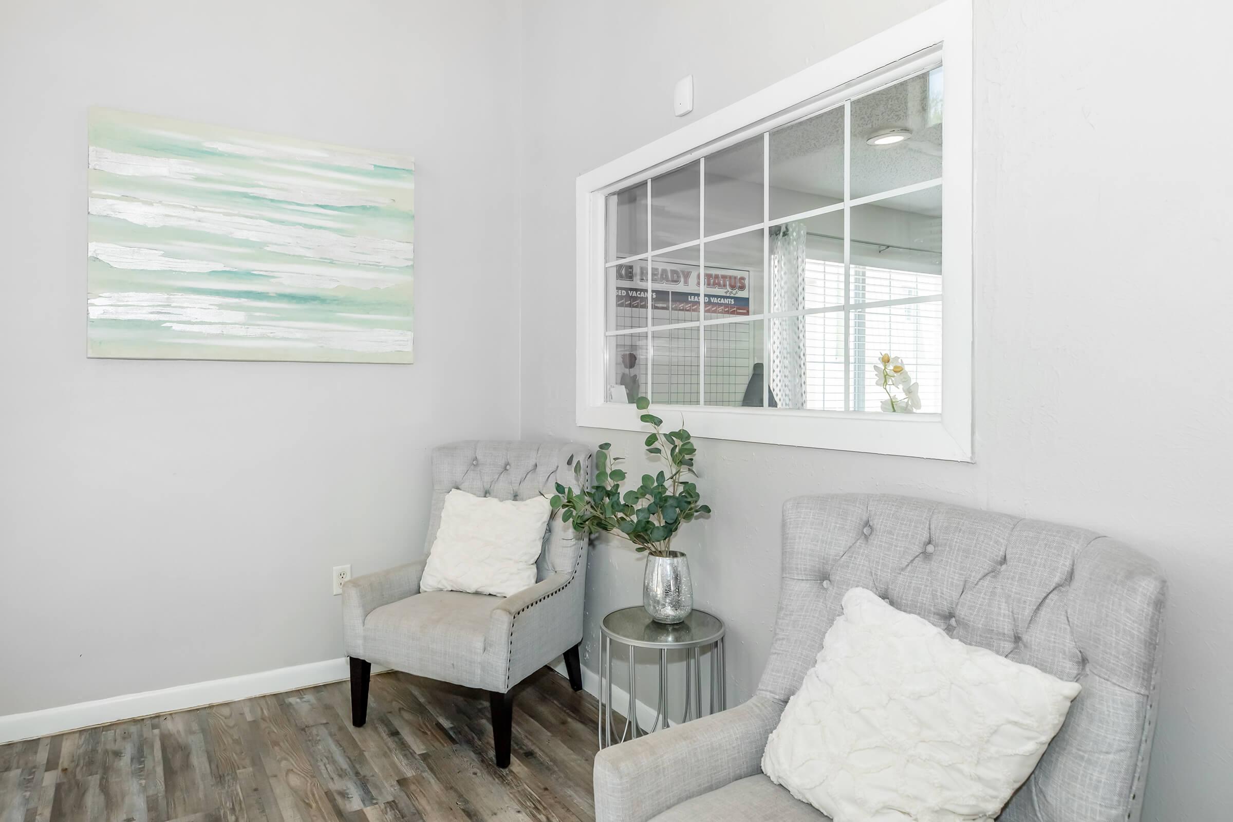 A cozy interior space featuring two tufted gray chairs with white throw pillows. A small round side table holds a vase with greenery. In the background, there is a large window with sheer curtains and a light-colored abstract painting on the wall, enhancing the bright and inviting atmosphere.