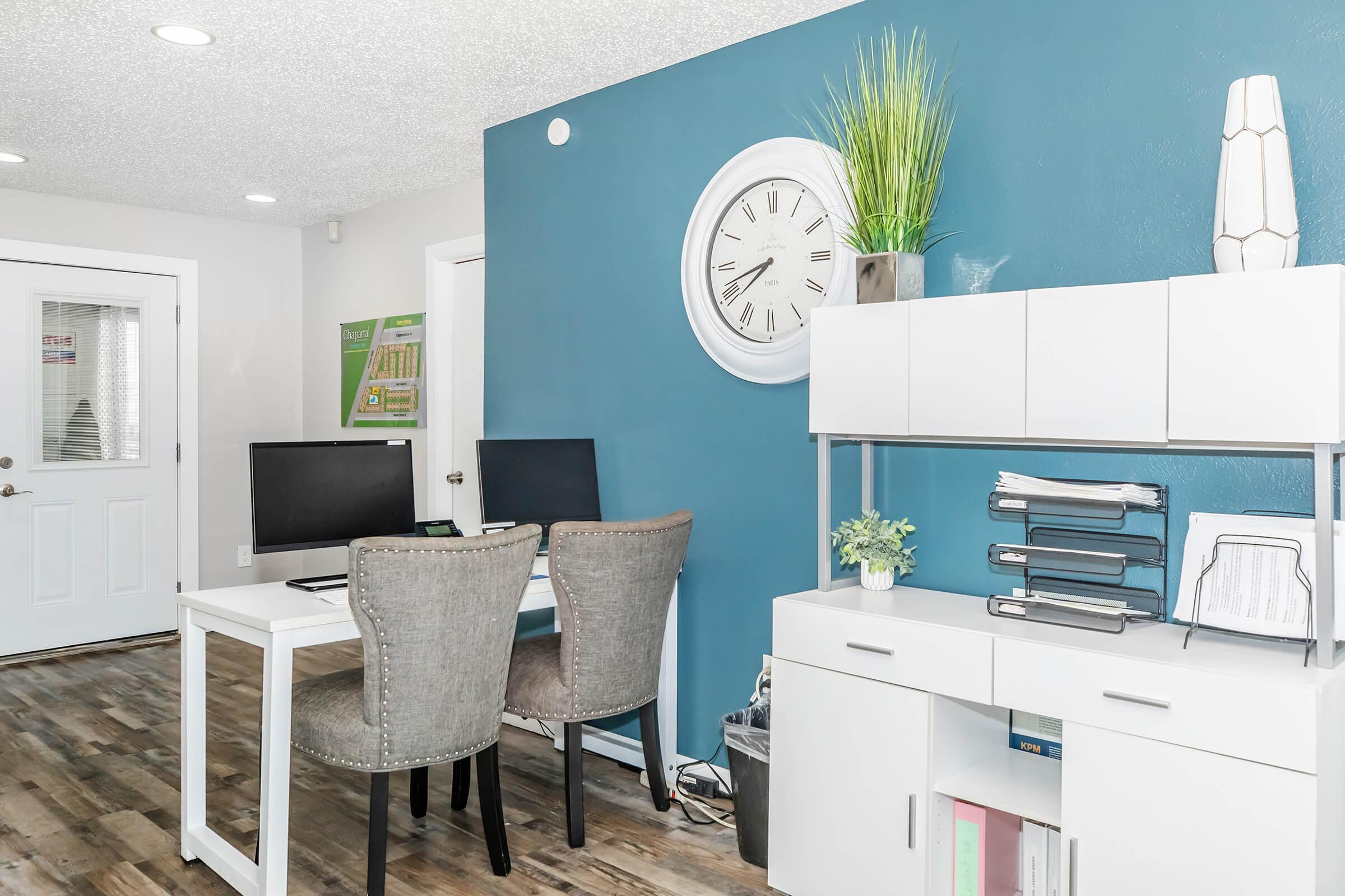 A modern office space featuring two computer desks with gray chairs, a white shelf with decorative items and books, and a large wall clock. The walls are painted a bright blue, and there is a door visible to the left, along with greenery decor in the room. The flooring is wood-look laminate.