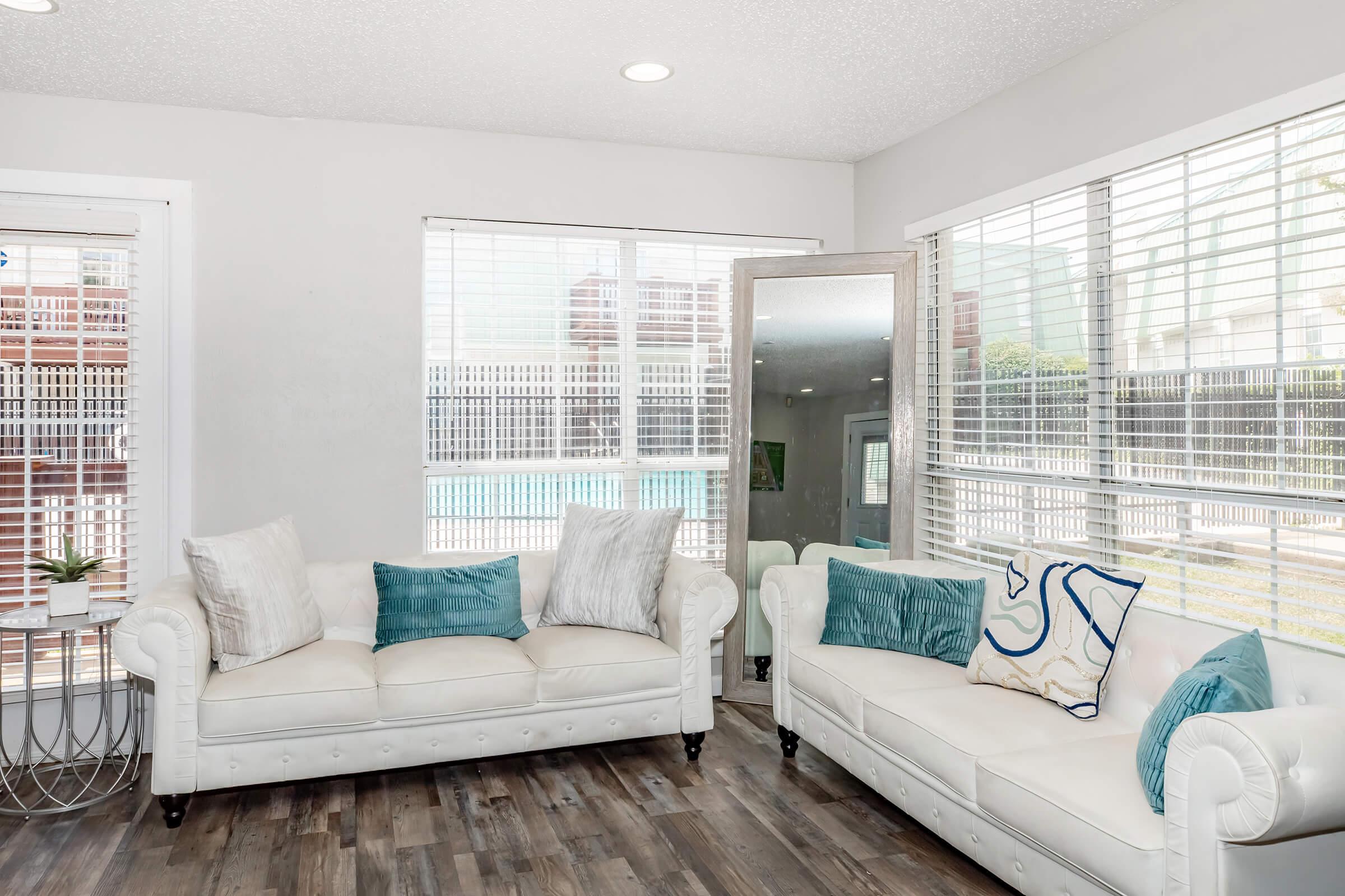 Bright and airy living room featuring two white sofas with teal and patterned throw pillows. Large windows allow natural light and provide a view of an outdoor pool area. The floor is made of wooden planks, and a small plant is placed on a round table near the sofas.