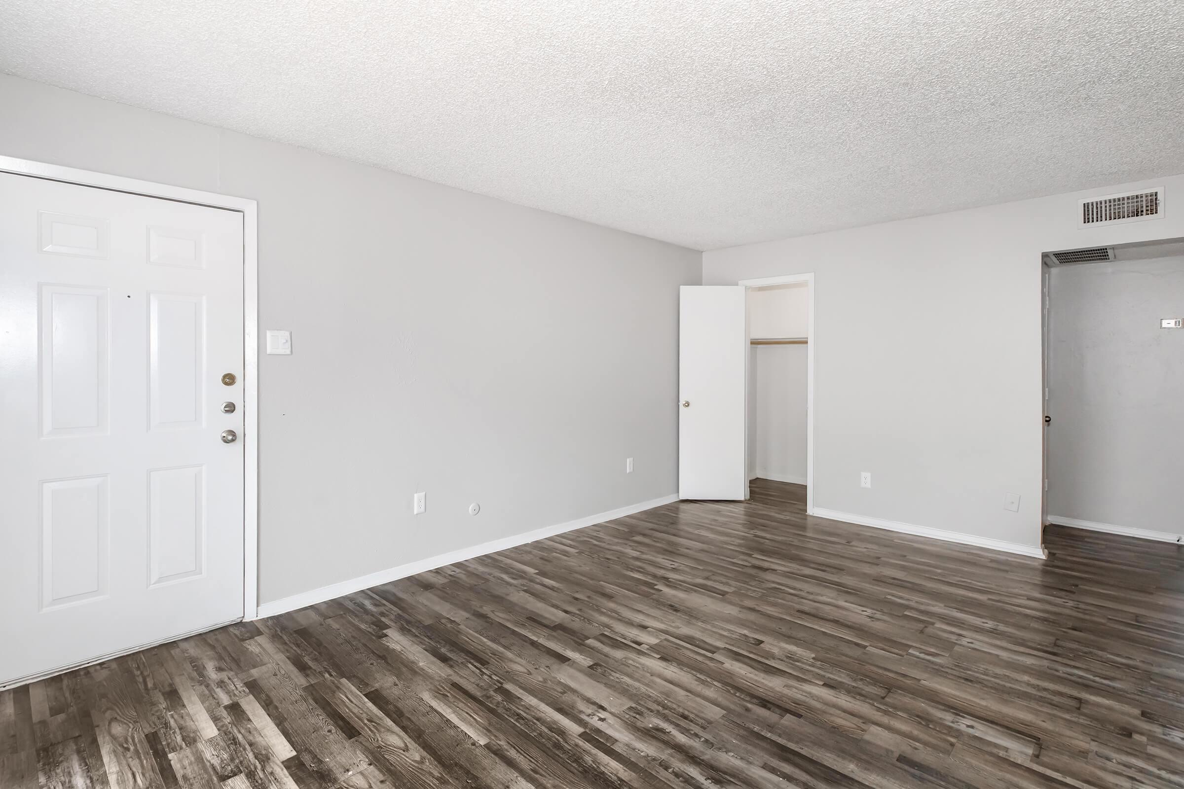 A spacious, light-filled living area featuring a textured ceiling, light gray walls, and modern laminate flooring. The entrance door is on the left, with a closet door visible in the background. The room is empty, creating a blank canvas for potential furniture arrangements.