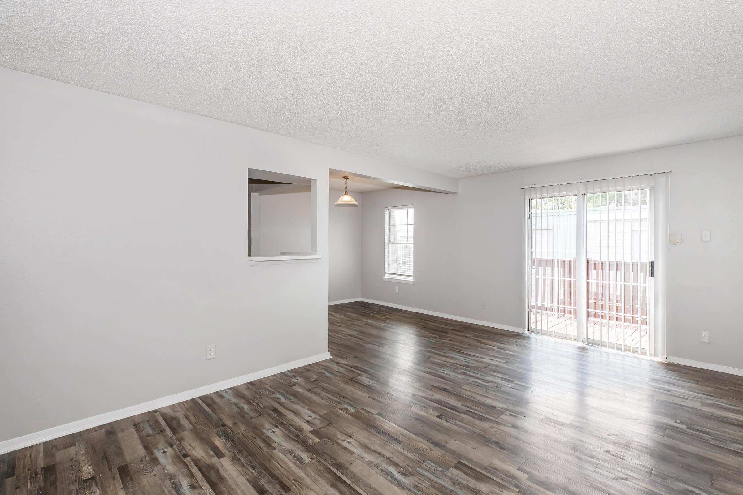 Spacious living room with gray walls and wooden flooring, featuring a light fixture hanging from the ceiling. There is a small opening to an adjacent room and a sliding glass door leading to an outdoor area, providing natural light and an inviting atmosphere.