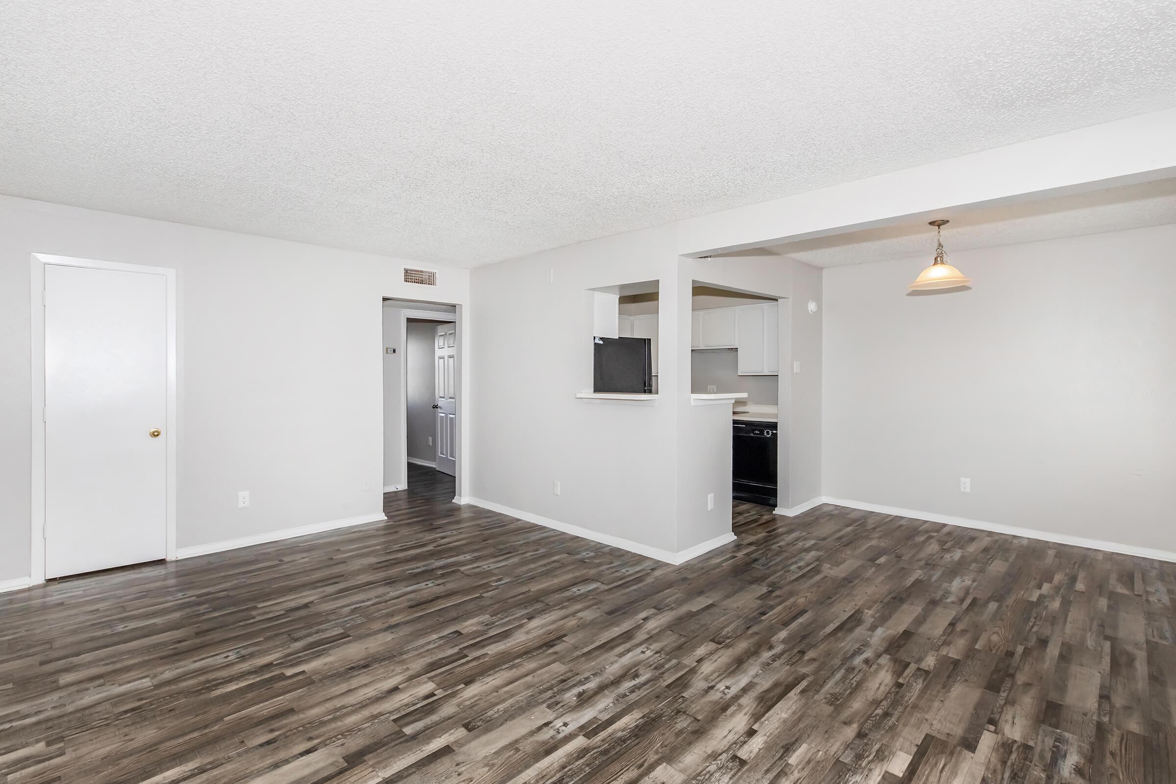 Interior view of an empty living space with a light wooden floor, light gray walls, and natural lighting. The area features a small kitchen visible in the background, a doorway leading to another room, and a pendant light hanging from the ceiling. The space appears clean and modern, ready for furnishing.