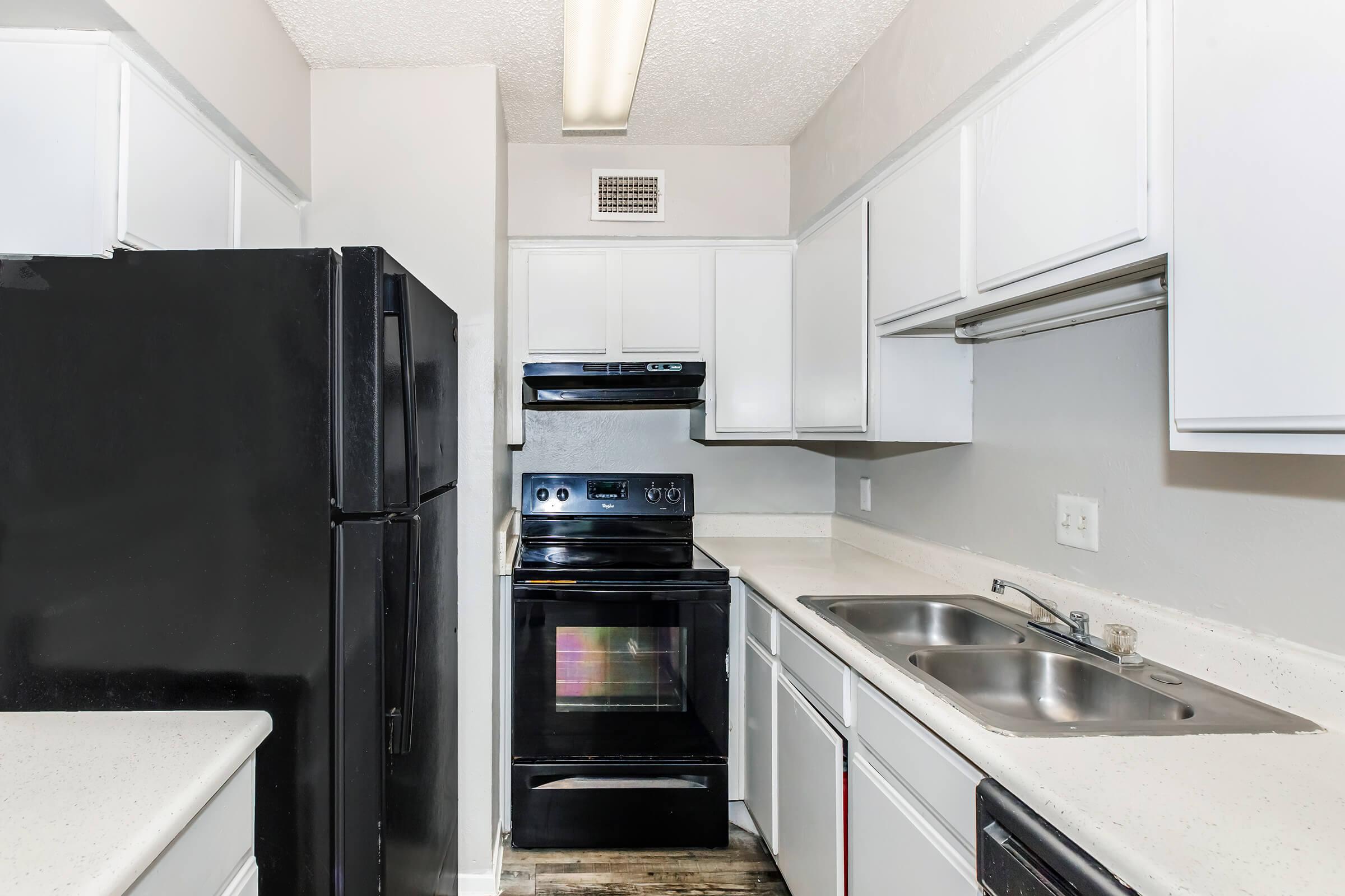 A modern kitchen featuring white cabinetry, a black refrigerator, an oven, and a stovetop. The countertops are light-colored, and there are double sinks. The space is well-lit with overhead lighting and has a neutral wall color. The layout is compact and functional.