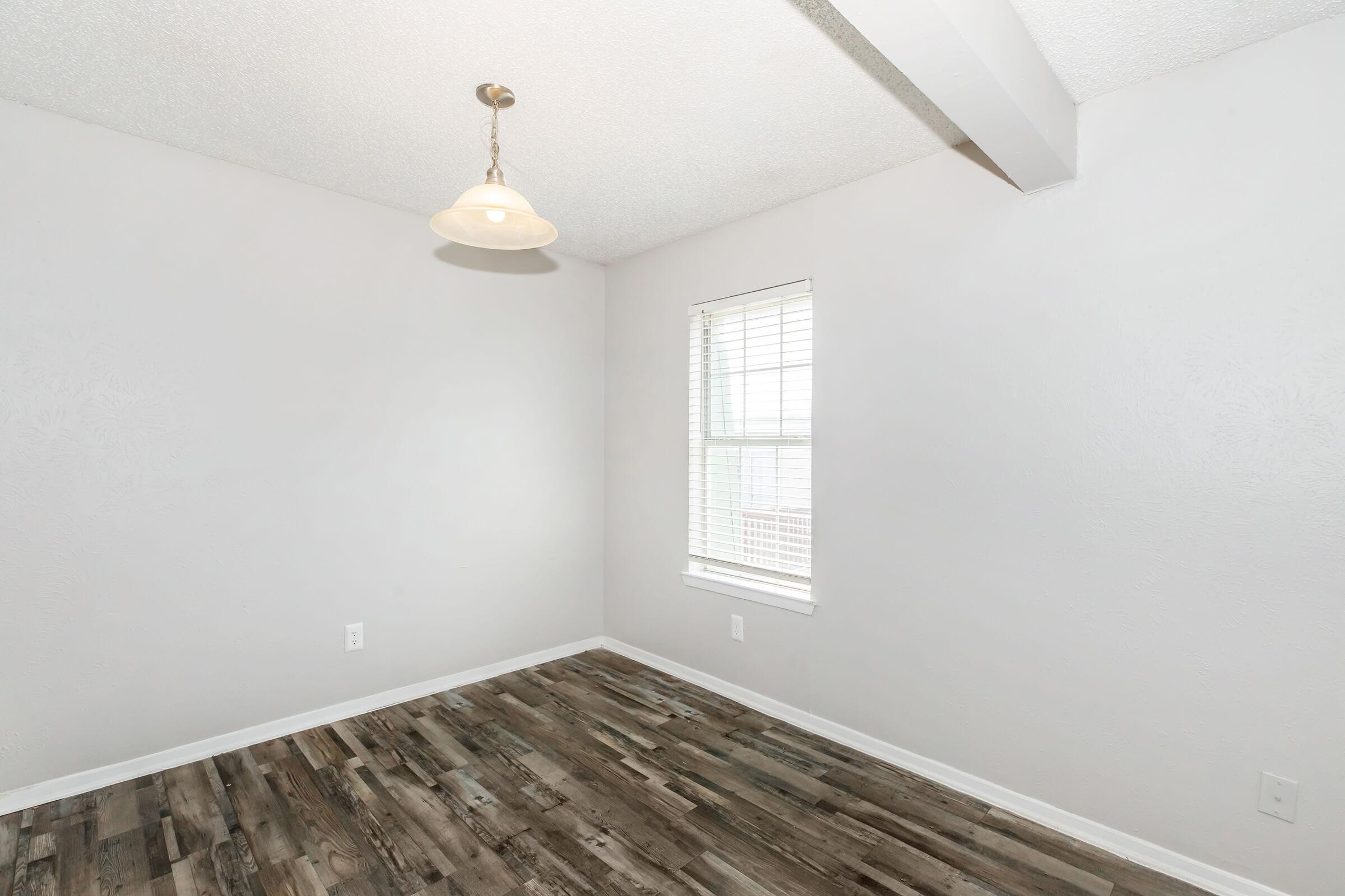 A bright, empty room featuring light-colored walls and a wooden floor. There's a small window with blinds allowing natural light in, and a hanging light fixture on the ceiling. The room has a simple and clean design, ideal for various uses.