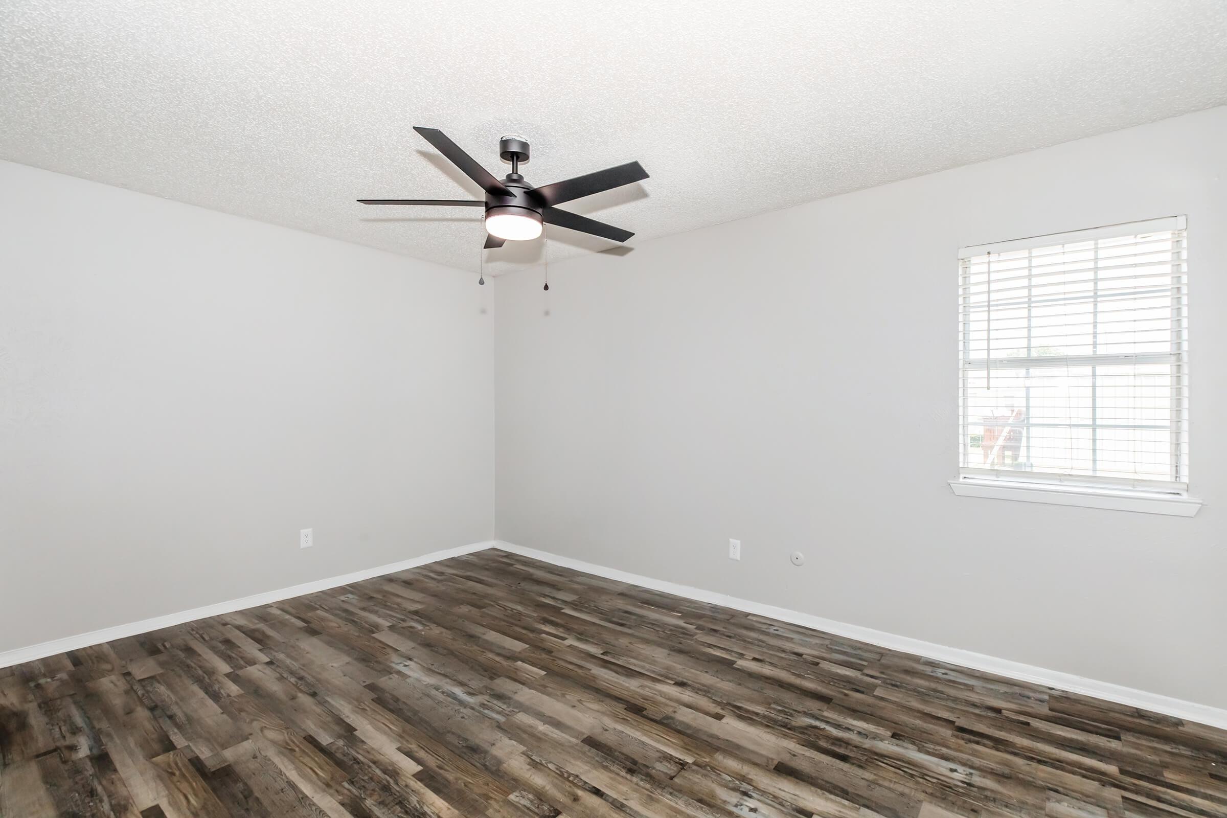 Empty room with gray walls and a ceiling fan. The floor features wood-like planks, and there is a window with light-colored blinds letting in natural light. The room is spacious and unobstructed, showcasing a clean and modern design.