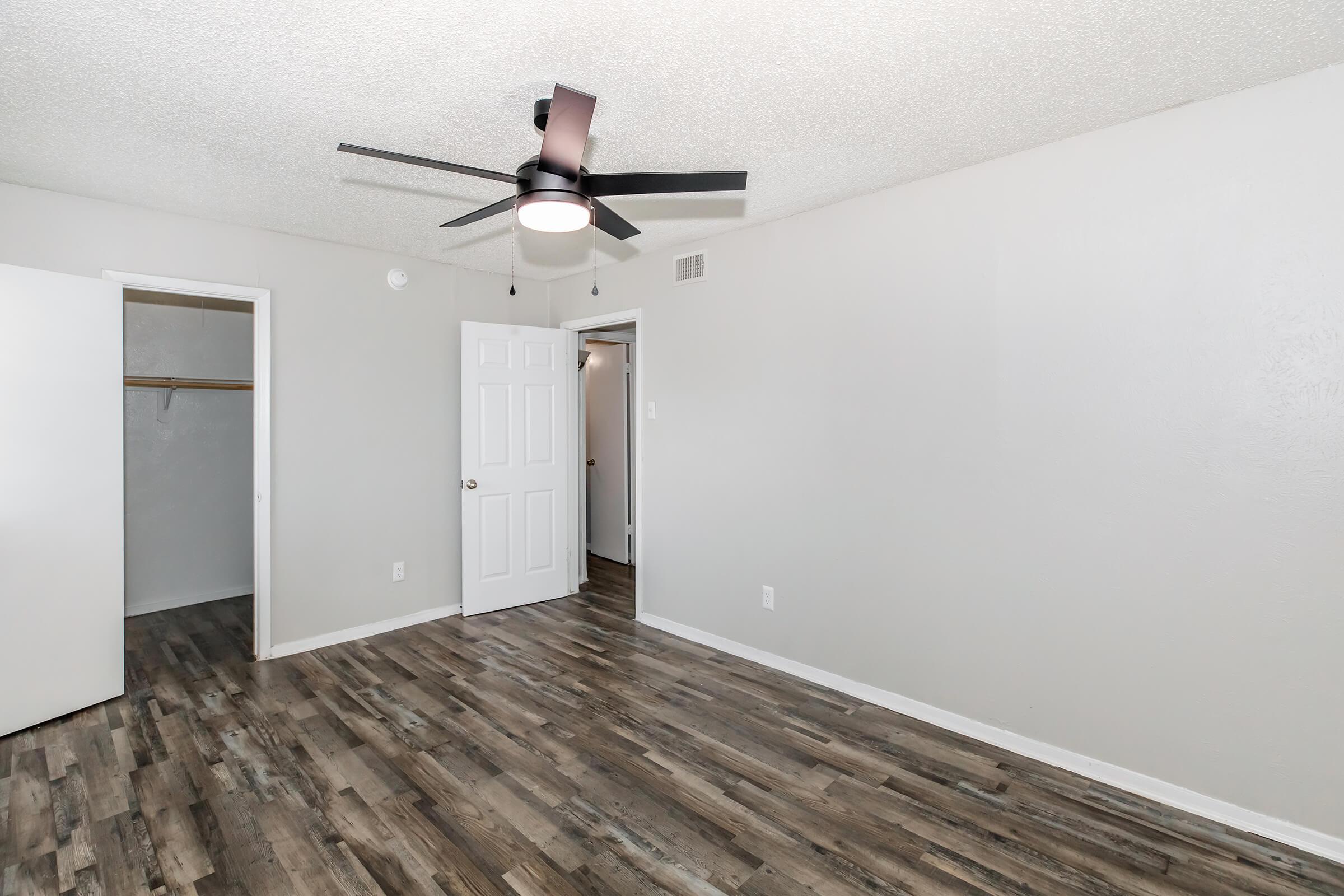 Interior of a room featuring a ceiling fan, light gray walls, and wood-like flooring. A white door leads to a closet on the left, while another door on the right opens to a hallway. The natural light illuminates the space, creating a bright and airy atmosphere.