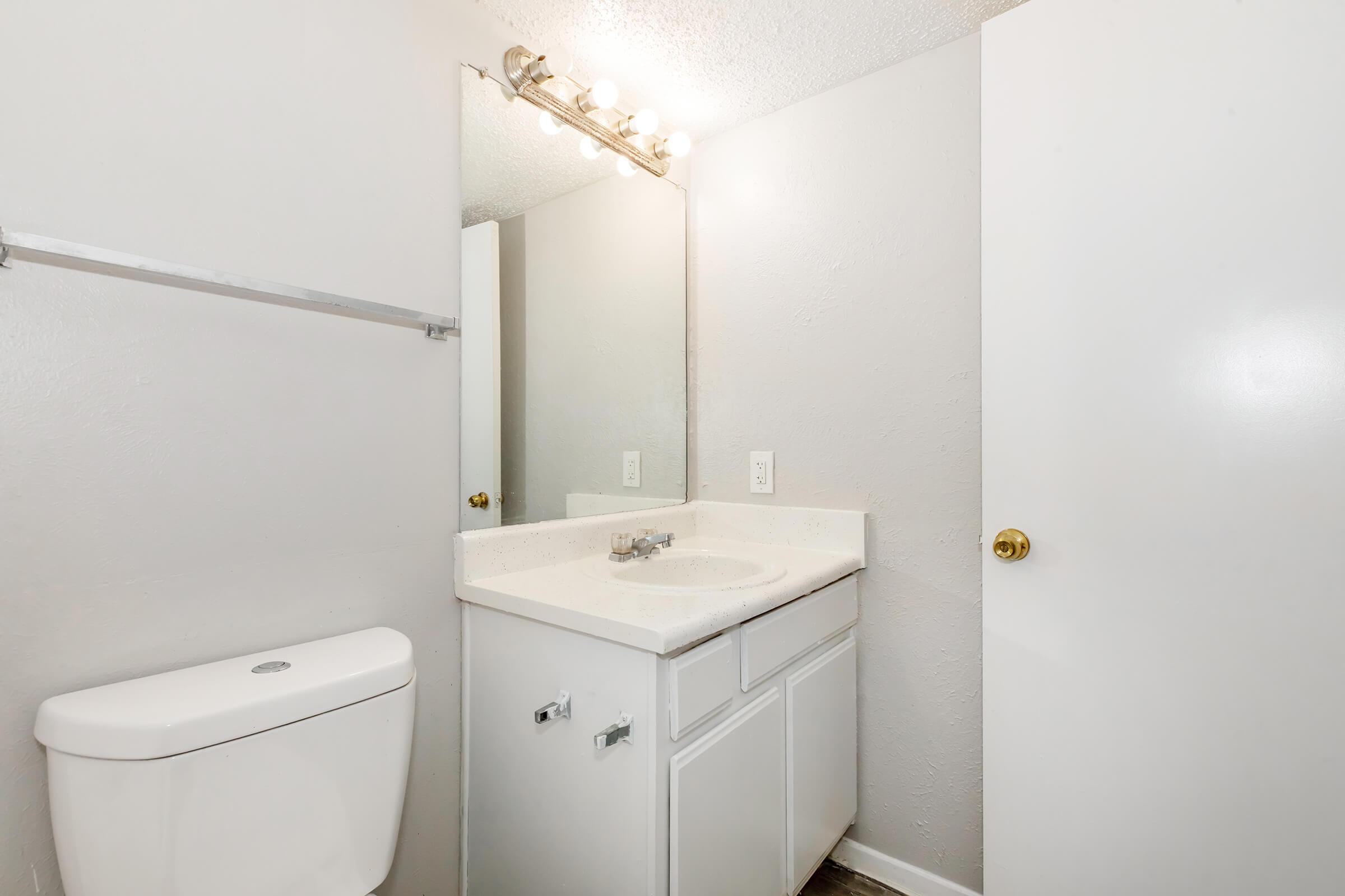 A simple bathroom featuring a white toilet, a sink with a countertop, a large mirror above the sink, and a bright light fixture. The walls are painted grey, and there is a cabinet under the sink. A white door is partially closed, adding to the minimalistic design.
