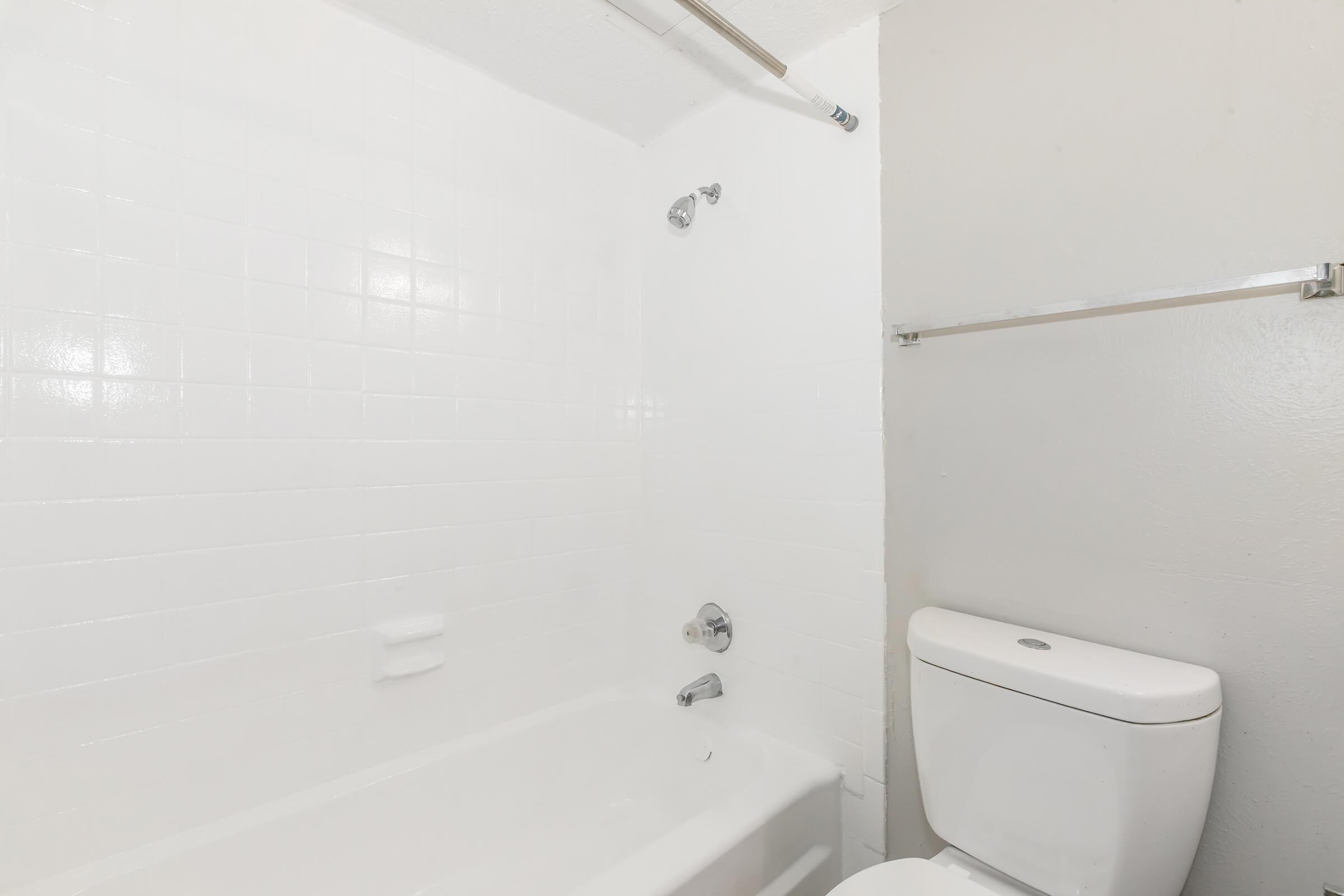 A clean, minimalistic bathroom featuring a white bathtub with a showerhead, a wall-mounted grab bar, and a white toilet. The walls are tiled in white, creating a bright and spacious feel, with a simple, light-colored wall on the side.