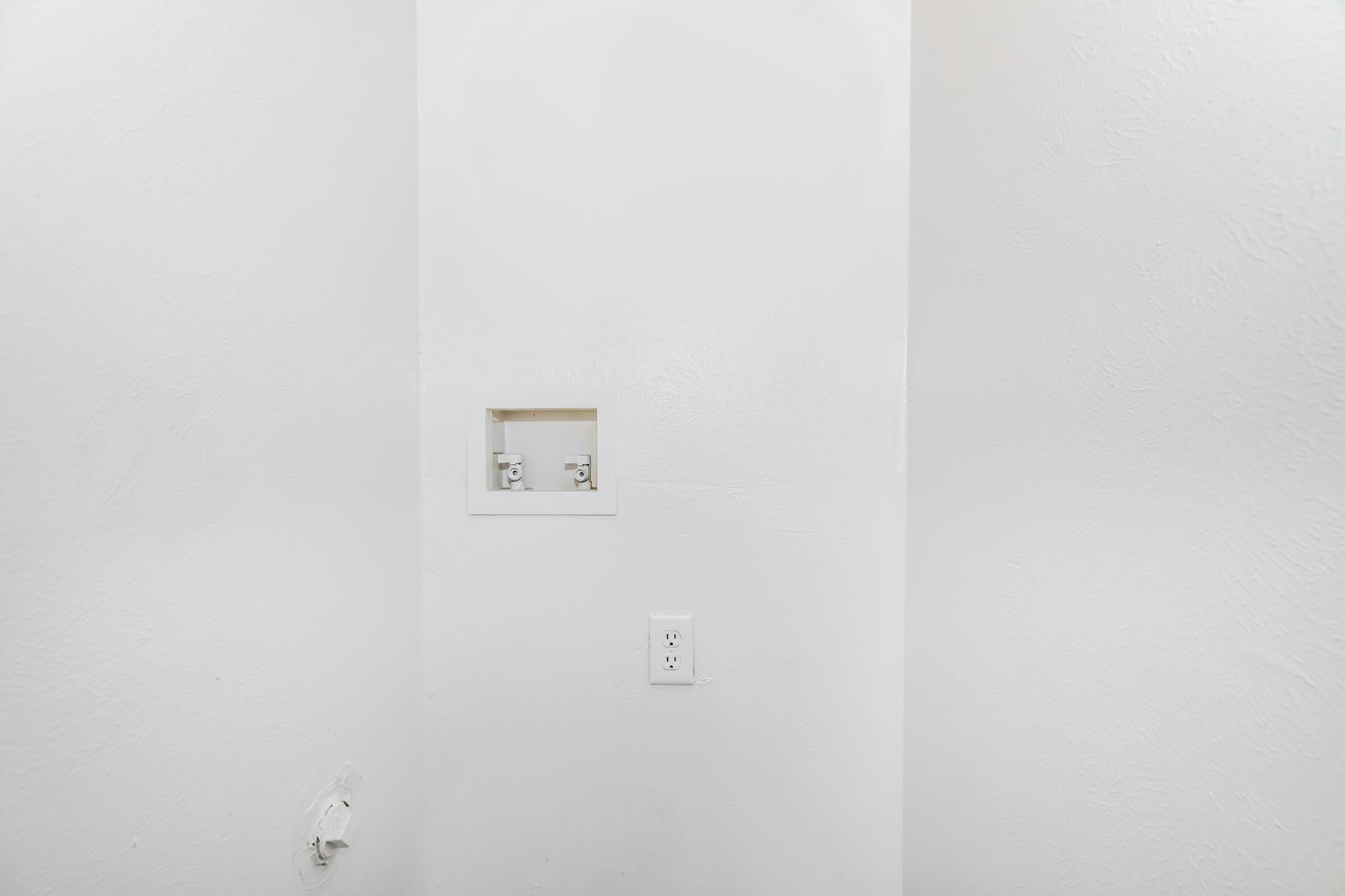 A mostly empty white wall with a small recess containing two electrical outlets. To the right, there is a visible power outlet. The overall appearance is clean and minimalistic, with no additional decorations or furniture in view.