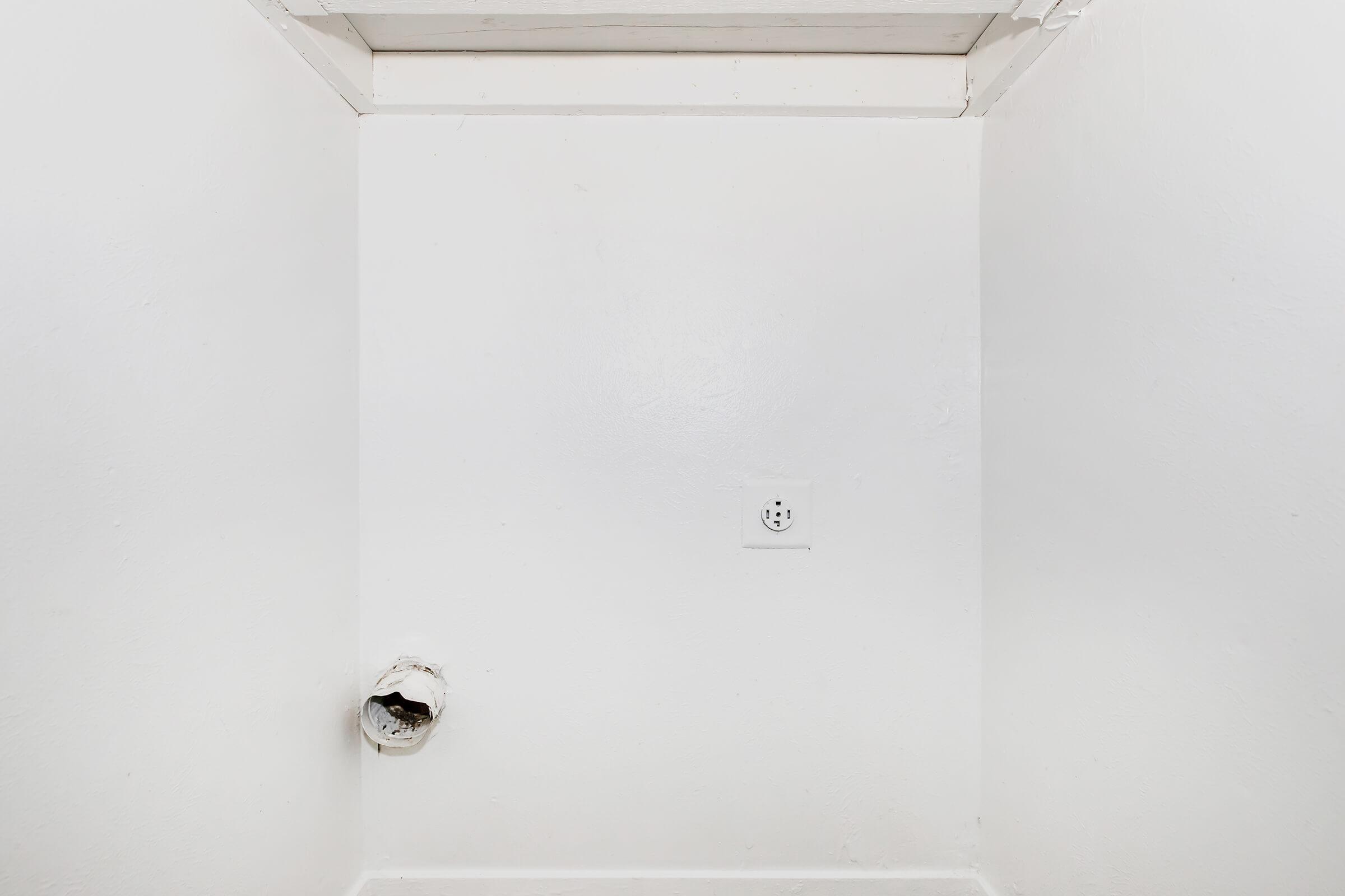 A plain white wall with a small circular ventilation duct on the left and an electrical outlet on the right. The surface appears smooth and well-maintained, with no additional features or decorations. The overall scene has a minimalist aesthetic.