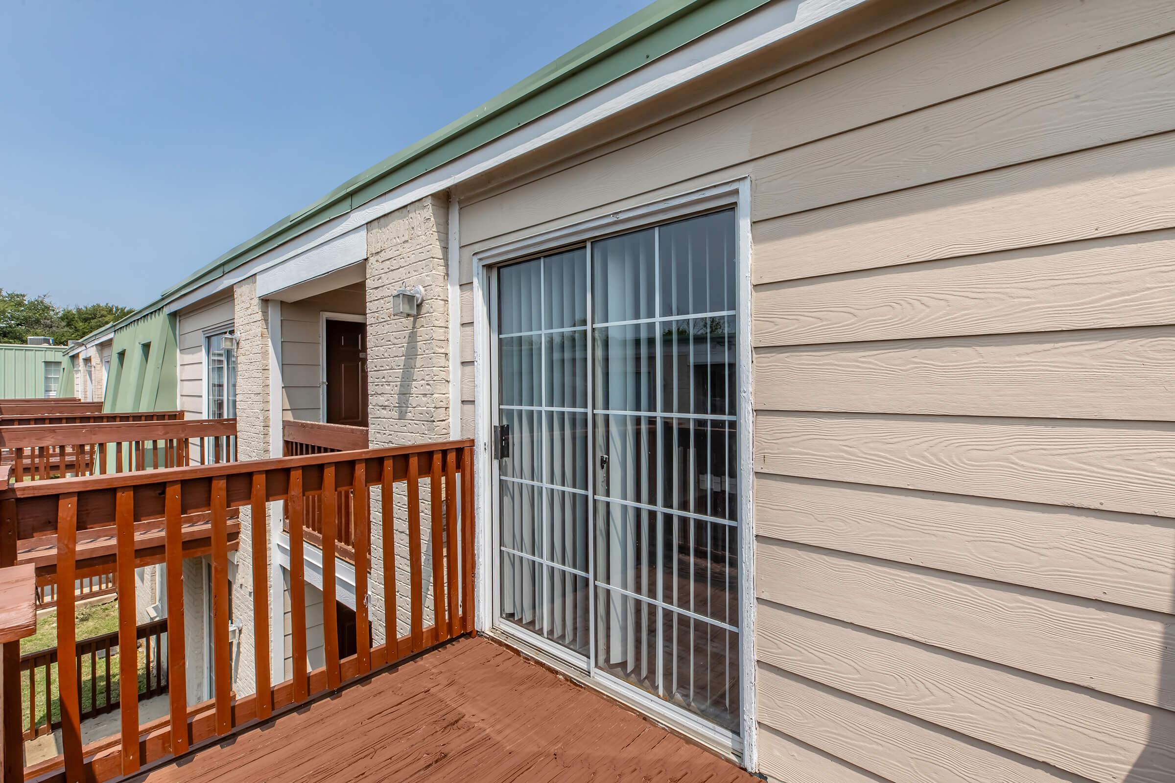 A view of a wooden balcony with a sliding glass door leading into a room. The exterior walls are light-colored, and the building features a green roof. The balcony has a wooden railing, and there is a light fixture beside the door. A grassy area is visible in the background.