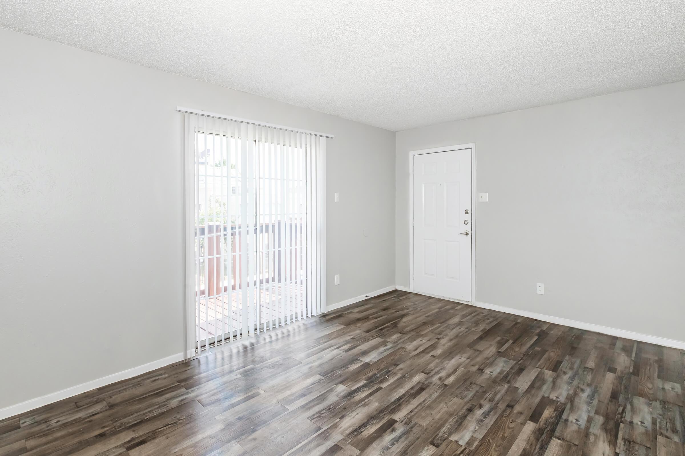 Empty living room with light gray walls and a textured ceiling. Features a sliding glass door with vertical blinds leading to an outdoor area. The flooring is a dark wood laminate. A plain white door is visible on the right side, and the space is well-lit with natural light.