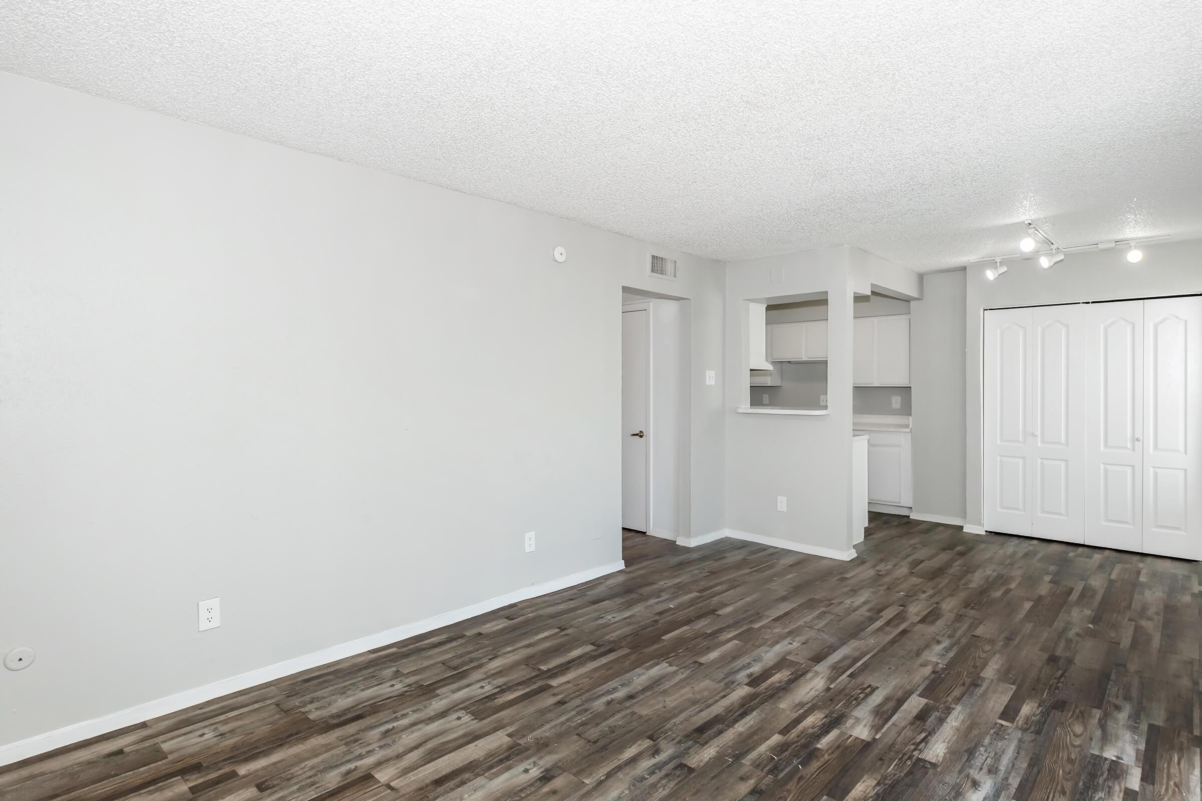 An interior view of an apartment featuring light gray walls, a textured ceiling, and a wooden floor. There is a doorway on the left leading to another room, a compact kitchen area with a counter visible in the background, and a closet with sliding doors on the right. The space is well-lit and open.