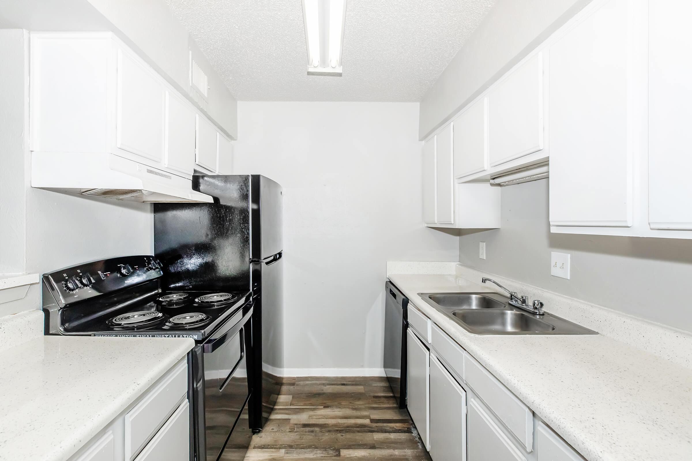 A modern kitchen featuring white cabinetry, a black stove with four burners, a black refrigerator, and a double sink. The countertops are light-colored and there is a light fixture overhead. The walls are painted in a neutral tone, and the floor has a wood-like finish.