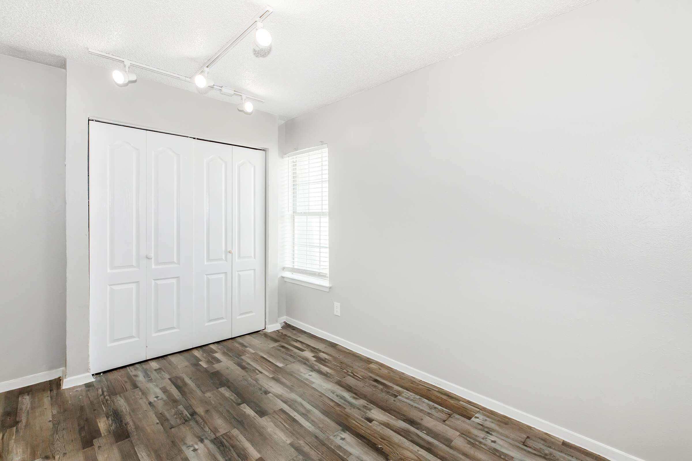 A clean, empty room with light gray walls and wooden flooring. There's a white closet with double doors on one side and a window with blinds allowing natural light. The ceiling has track lighting, creating a modern and bright atmosphere.