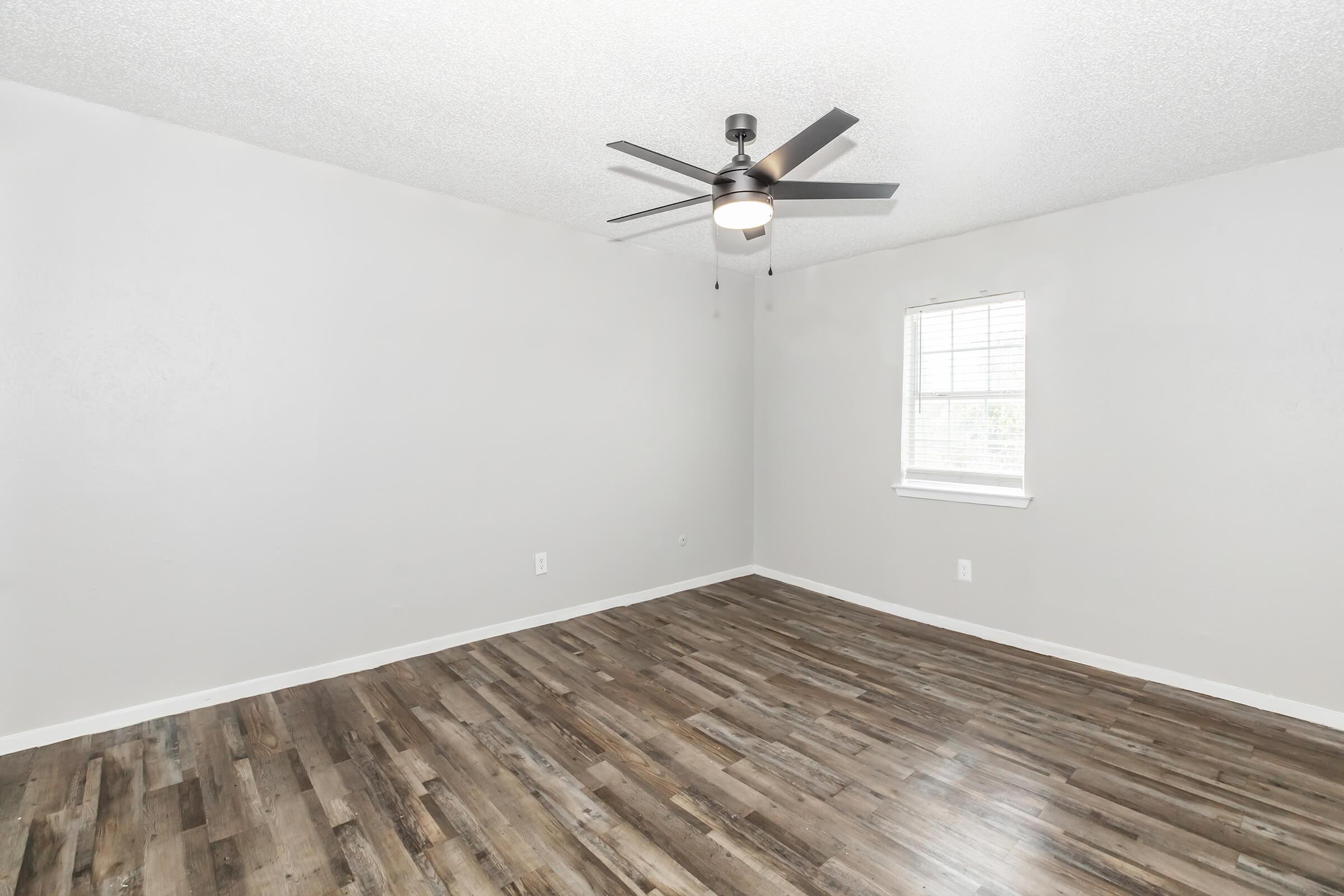 A spacious, empty room with light gray walls and a ceiling fan. The floor features dark wood laminate. A single window allows natural light to enter, enhancing the room's brightness.
