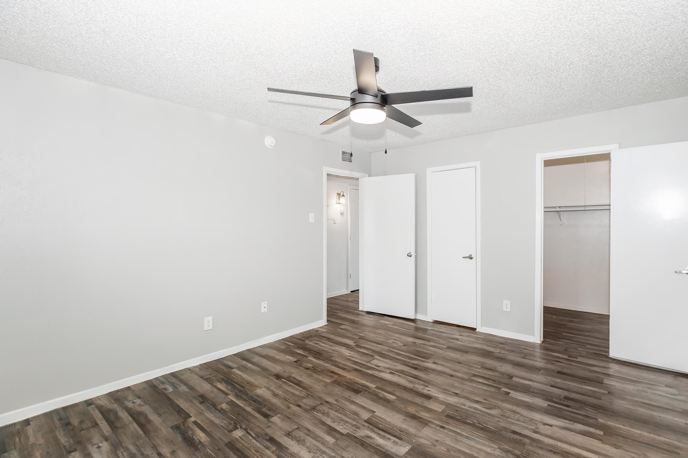 A spacious interior room featuring a ceiling fan, light gray walls, and laminate flooring. The room has two white closet doors and an open doorway leading to another area. The overall atmosphere is modern and inviting, with ample natural light.