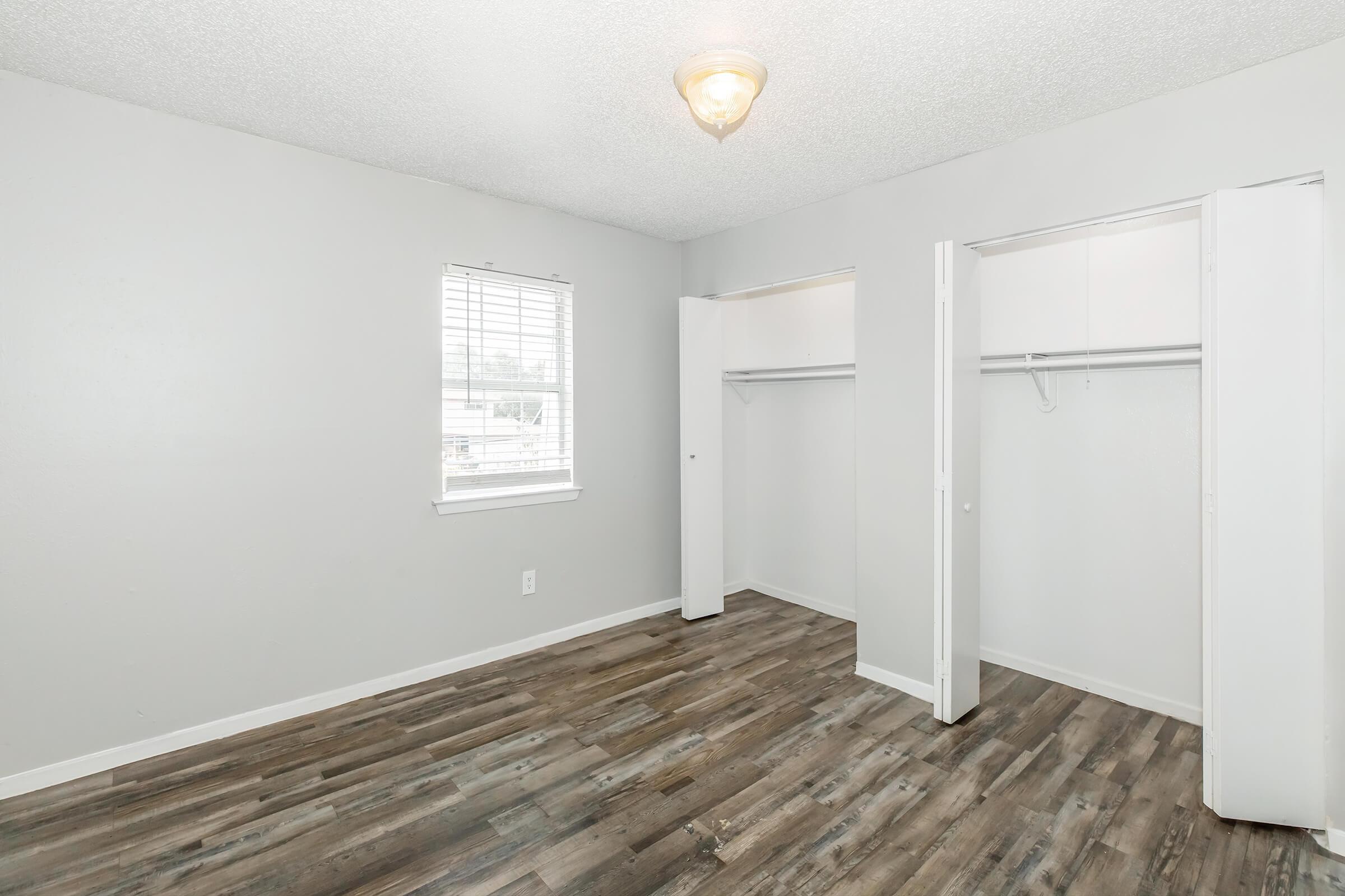 A small, empty bedroom with light gray walls and a window allowing natural light. The floor features wooden planks, and there are two open closets along one wall. A ceiling light fixture is visible, illuminating the space.