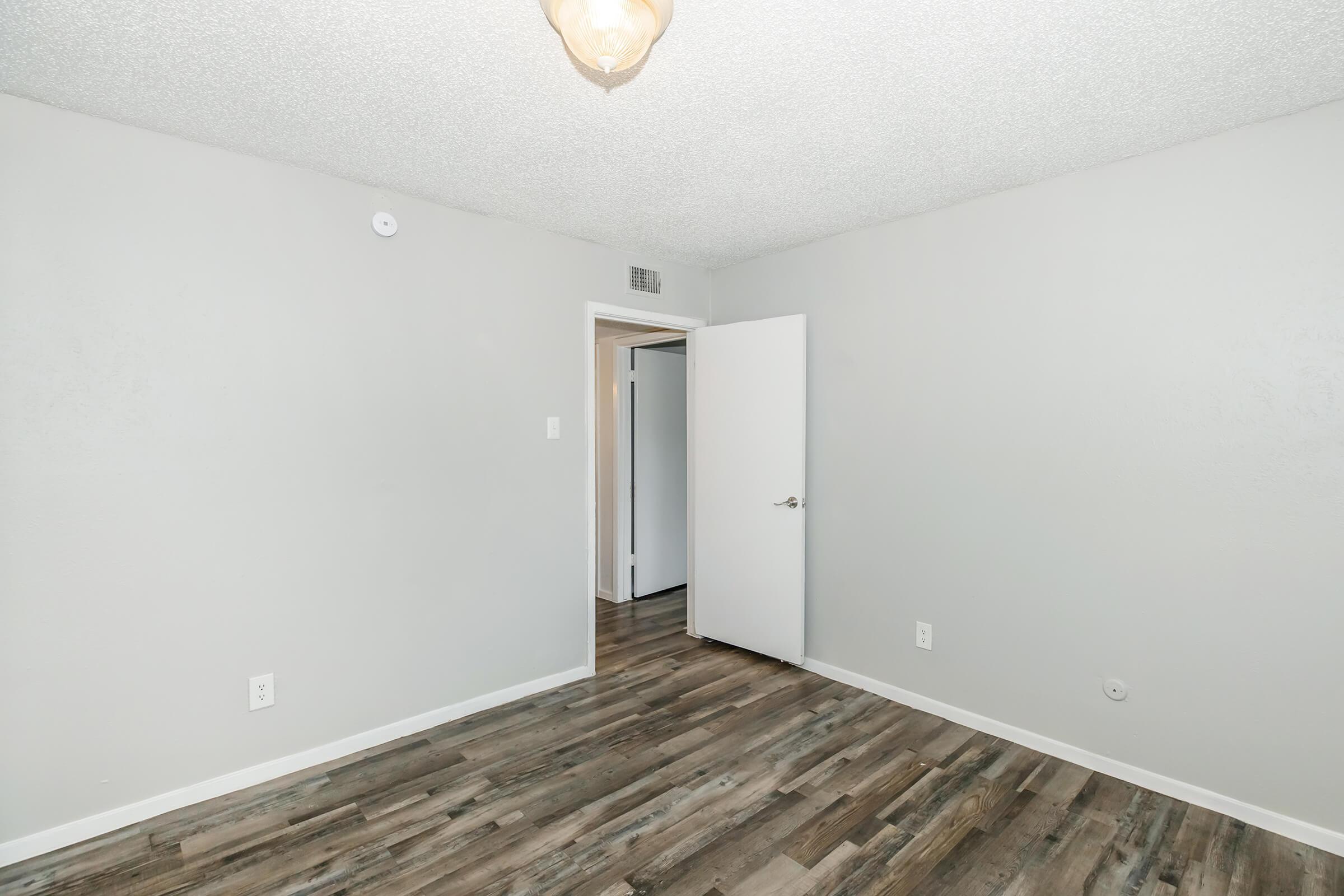 A simple, empty room with light gray walls and a textured ceiling. The floor has a wood-like finish, and there is a door on the left leading to another area. A light fixture hangs from the ceiling, providing soft illumination. The overall atmosphere is clean and minimalistic.