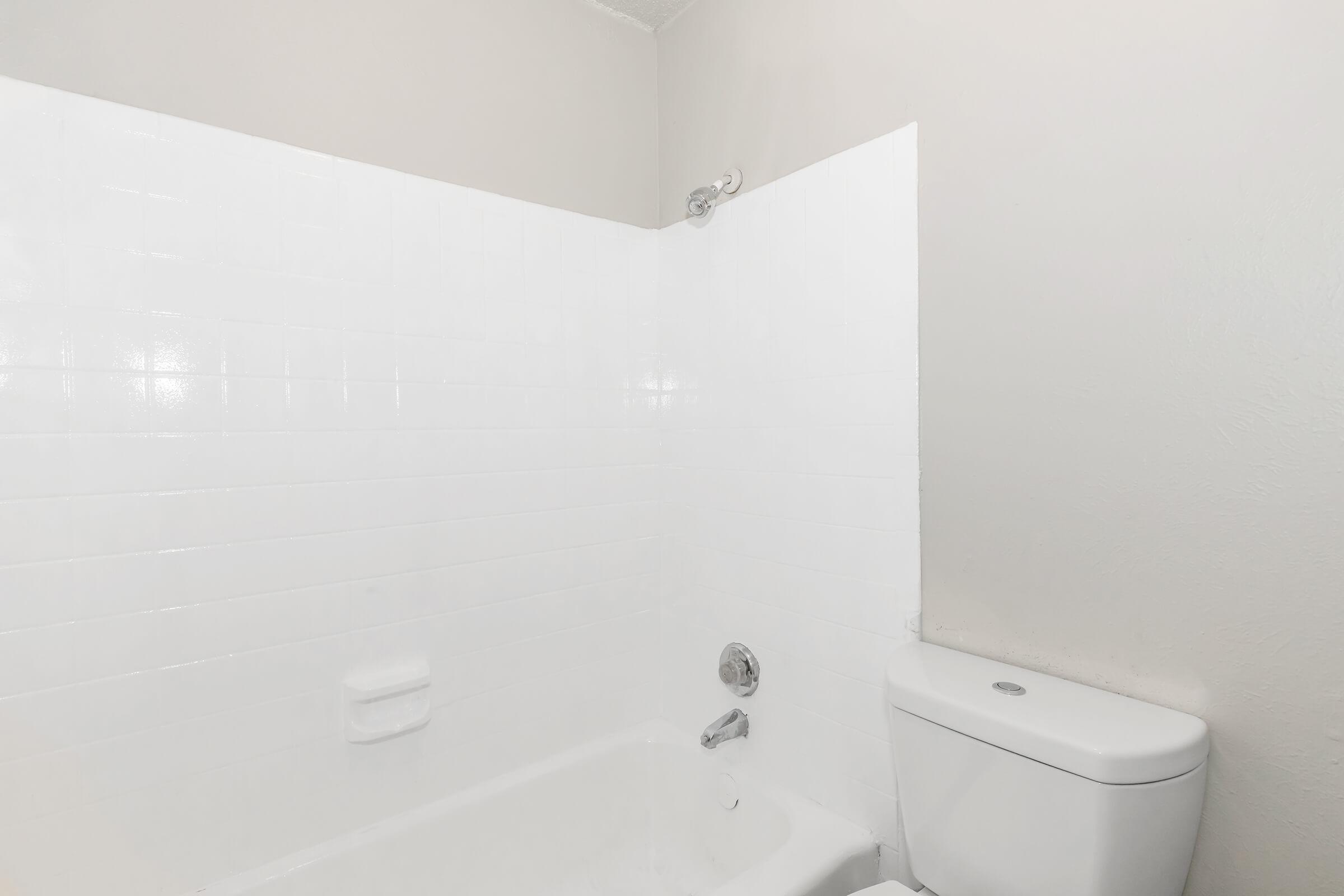 A clean and minimalistic bathroom featuring a white tiled shower area, a bathtub, and a toilet. The walls are painted in a light neutral color, enhancing the bright and open feel of the space.