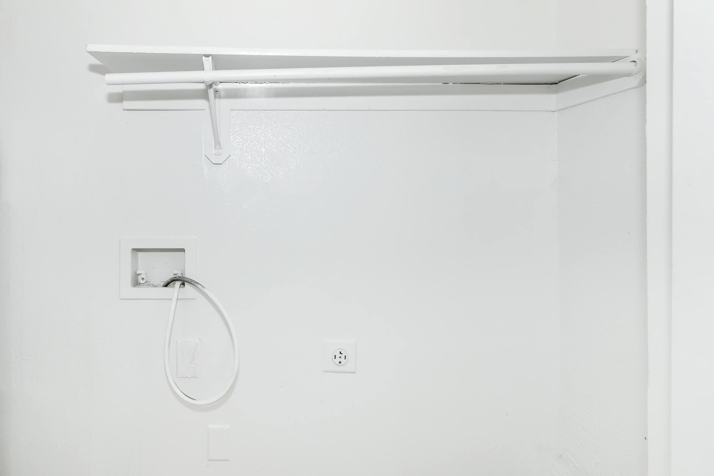 Empty white wall with a mounted shelf and a plumbing fixture. A flexible hose is connected to the fixture, and an electrical outlet is visible below. The overall appearance is minimalistic and clean, with a focus on the white surfaces and simple design elements.