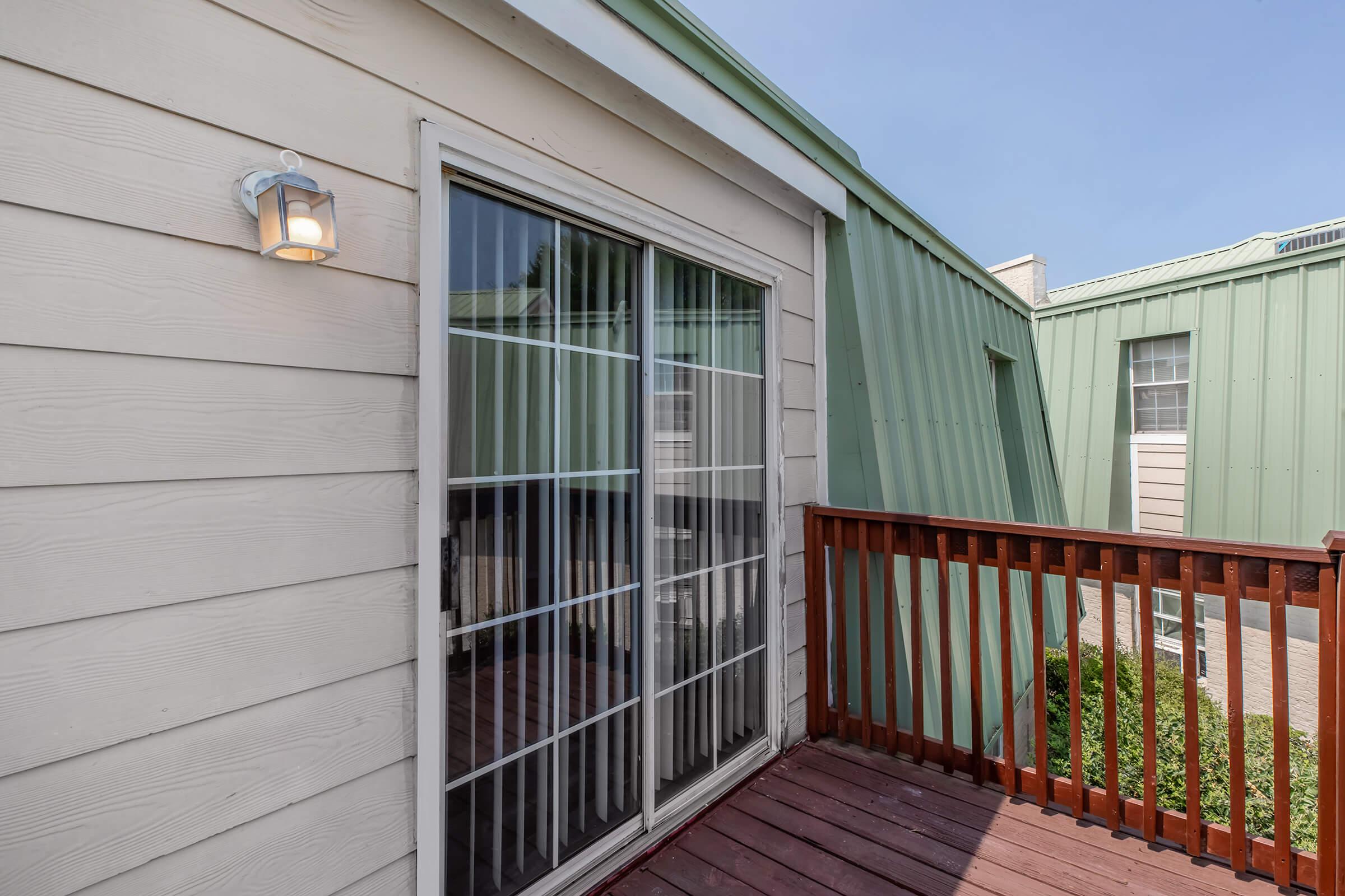 A balcony view featuring a sliding glass door framed by light-colored wooden siding. The balcony is enclosed with wooden railing, and there are green metal rooftops visible in the background under a clear blue sky. A light fixture is mounted on the wall beside the door.