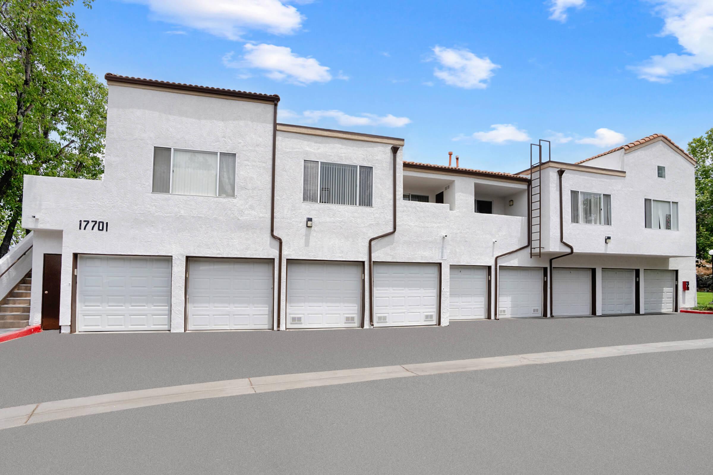 A modern two-story white building with multiple garage doors on the ground floor, situated in a landscaped area under a clear blue sky. The design features a mix of windows and balconies, enhancing its contemporary aesthetic.