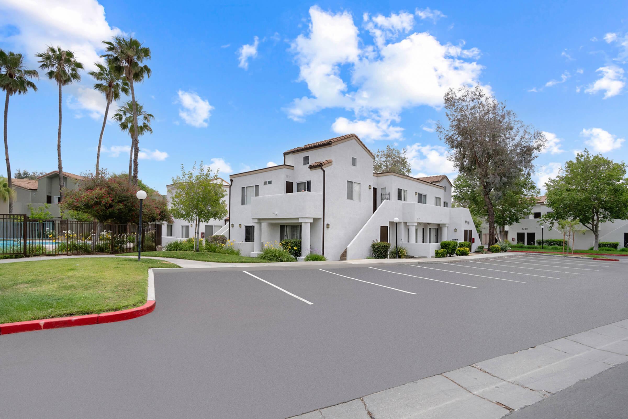 A modern multi-unit residential building surrounded by greenery, with palm trees in the background. In the foreground, there's a paved parking lot with several empty spaces. The sky is blue with a few clouds, creating a bright and inviting atmosphere.