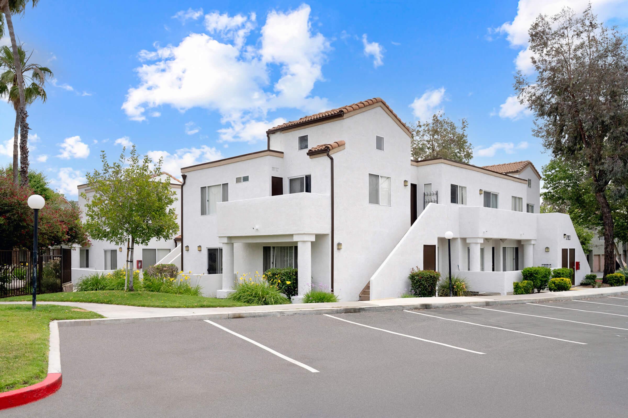 A modern, two-story residential building with white exterior and brown tile roofs, surrounded by green landscaping and palm trees. The parking area has several empty spaces, and the sky is partly cloudy. The entrance features a small staircase leading to balconies.