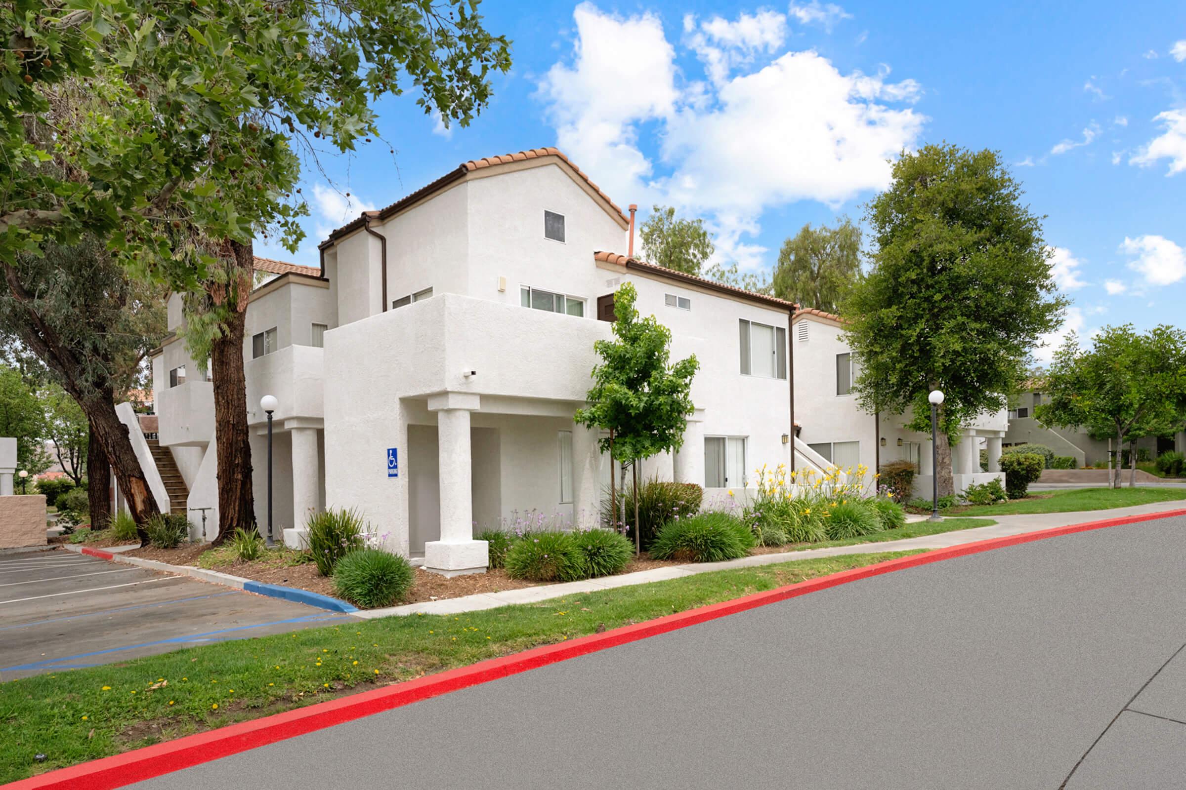 A modern residential building with multiple white stucco apartments surrounded by lush greenery. The scene features a clear blue sky with a few clouds, decorative yellow flowers, and a red-bordered path leading to the entrance. Parking spaces are visible nearby.