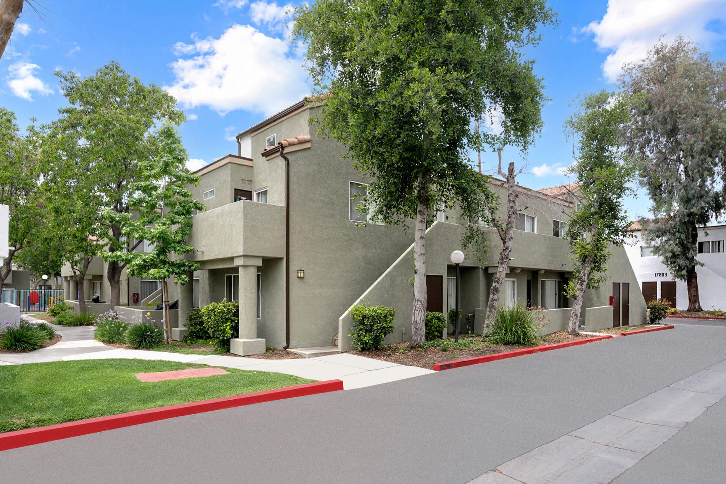 A peaceful residential area featuring modern two-story apartment buildings surrounded by trees and greenery. The pathway is bordered by well-maintained landscaping, and the street is clean with a red curb. Blue skies and fluffy clouds create a serene atmosphere.