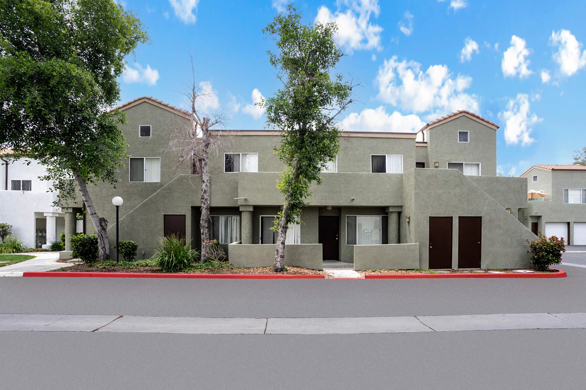 A modern, two-story apartment building with green exterior walls. The building features multiple windows and entrance doors, surrounded by small shrubs and trees. The sky is bright with fluffy clouds, and there is a paved path leading to the entrance, creating a welcoming atmosphere.