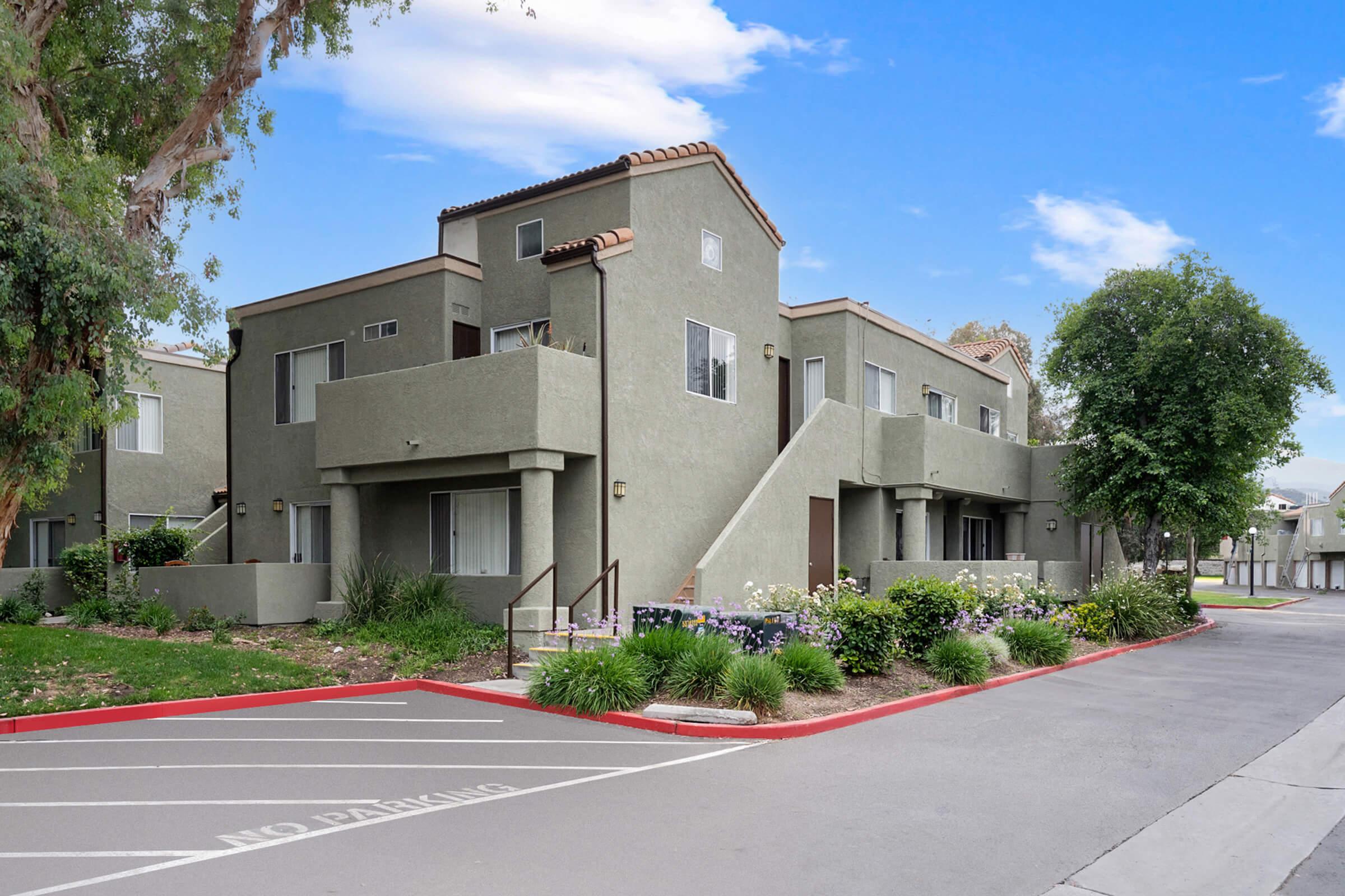 A multi-unit residential building with a light gray exterior. The structure features a tile roof, staircases leading to upper levels, and landscaped surroundings with shrubs and flowers. A "No Parking" sign is visible in the foreground, indicating designated parking areas. The sky is clear and blue.