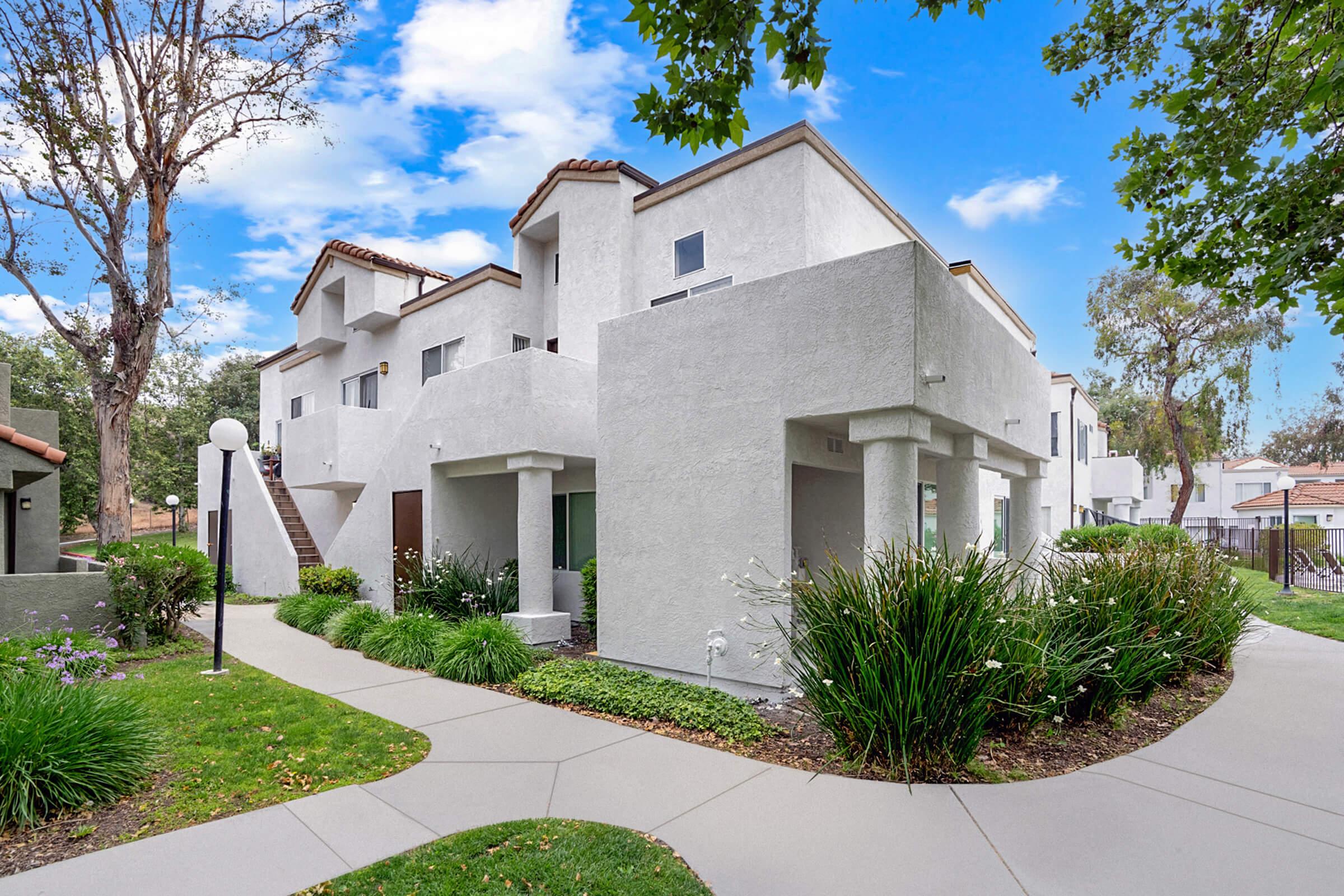 A modern, three-story white building with a stucco exterior, surrounded by greenery and well-maintained lawns. Pathways lead to the entrance, flanked by flower beds. Trees and blue skies are visible in the background, adding to a pleasant, residential atmosphere.