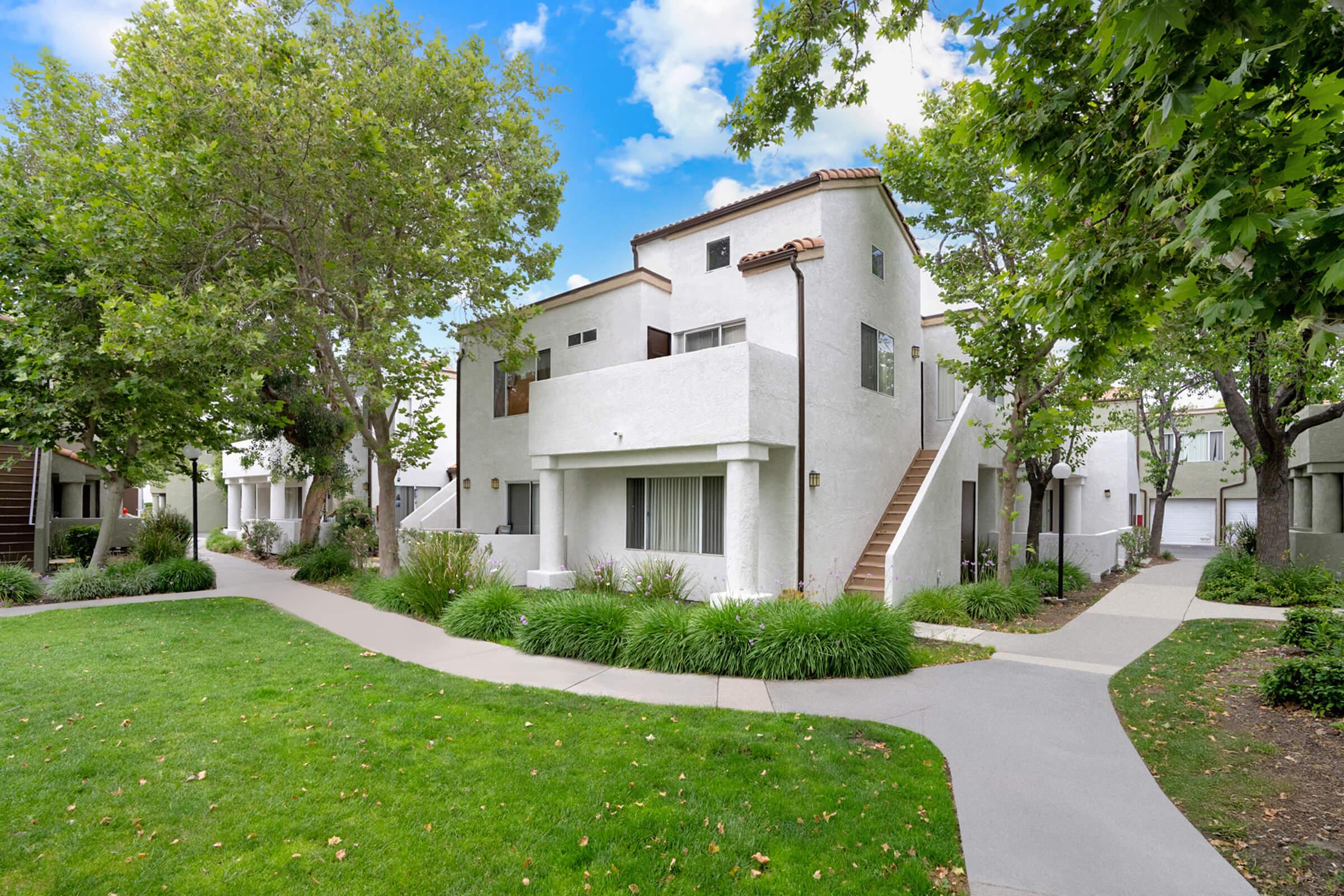 A modern two-story white building with a tiled roof, surrounded by greenery. There are trees and well-maintained lawns, with walkways leading to the entrance. The architecture features a blend of contemporary and traditional styles, set in a bright, sunny environment.