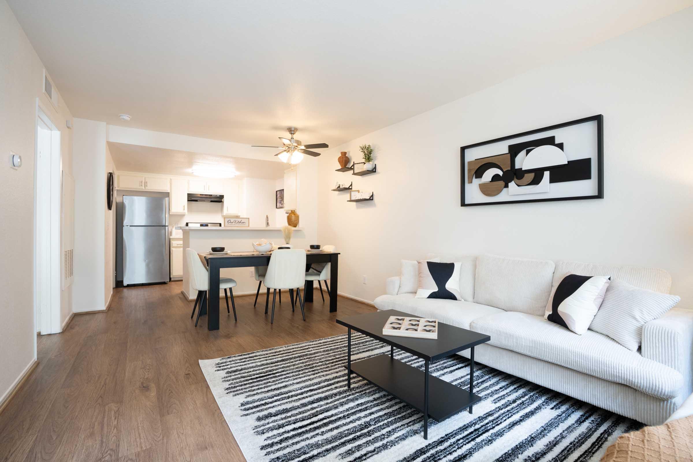 Modern living room and dining area featuring a white sofa, black coffee table, and a striped rug. A dining table with chairs is set beside a well-equipped kitchen with stainless steel appliances. The walls are adorned with minimalist art and decorative shelves. Bright, inviting atmosphere.