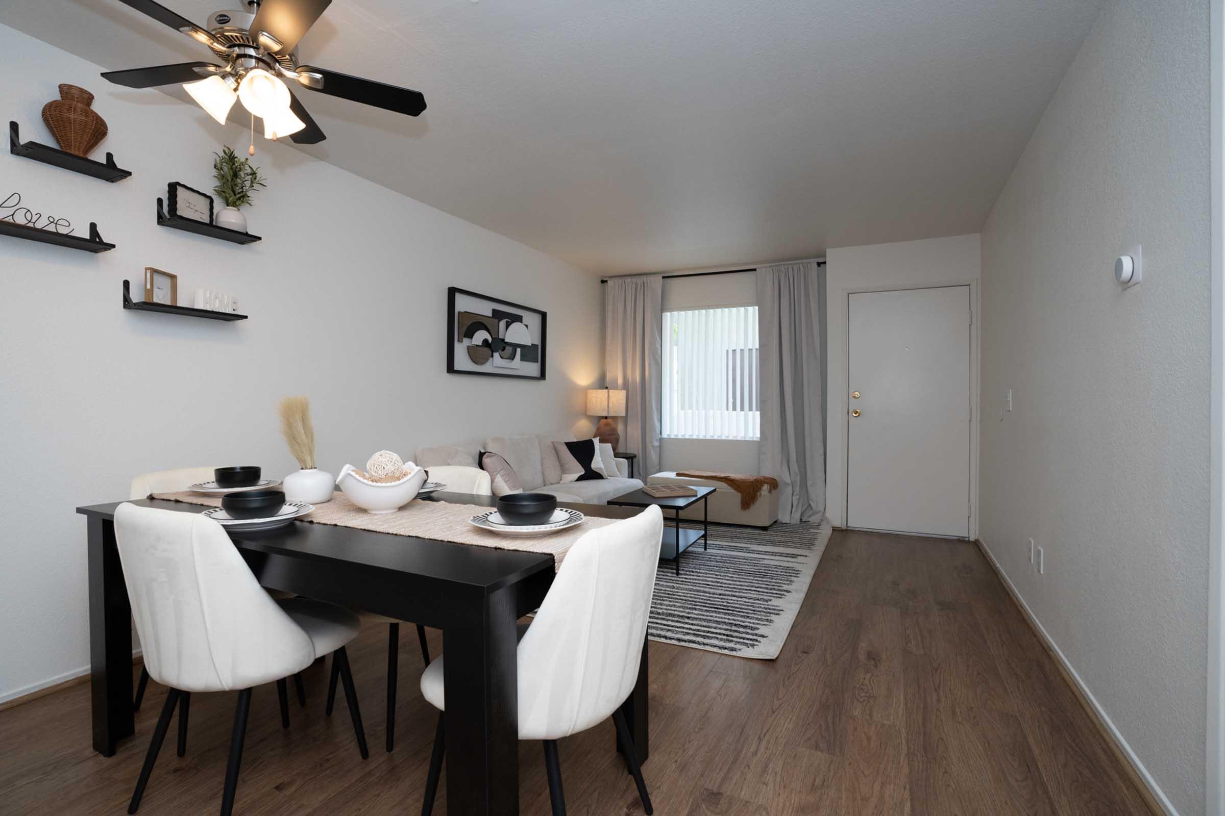 A modern living space features a dining table set for four with black and white tableware. The room has a stylish sofa, neutral decor, and a ceiling fan. A large window with light curtains allows natural light. Decorative shelves and a cozy area rug complete the inviting atmosphere.