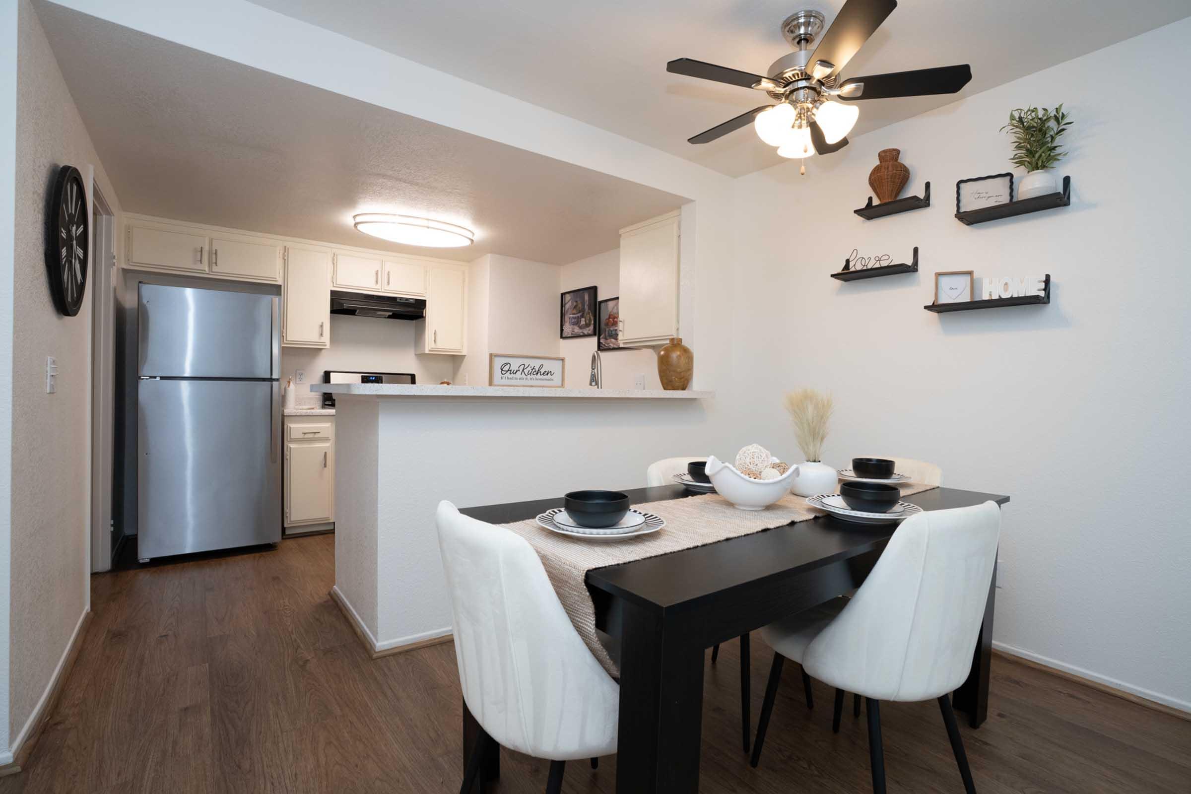 Modern kitchen and dining area featuring a black dining table with white chairs, set for a meal. The kitchen includes stainless steel appliances, a white countertop, and decorative items on the shelves. A ceiling fan provides light, and the space is bright and inviting with neutral colors.