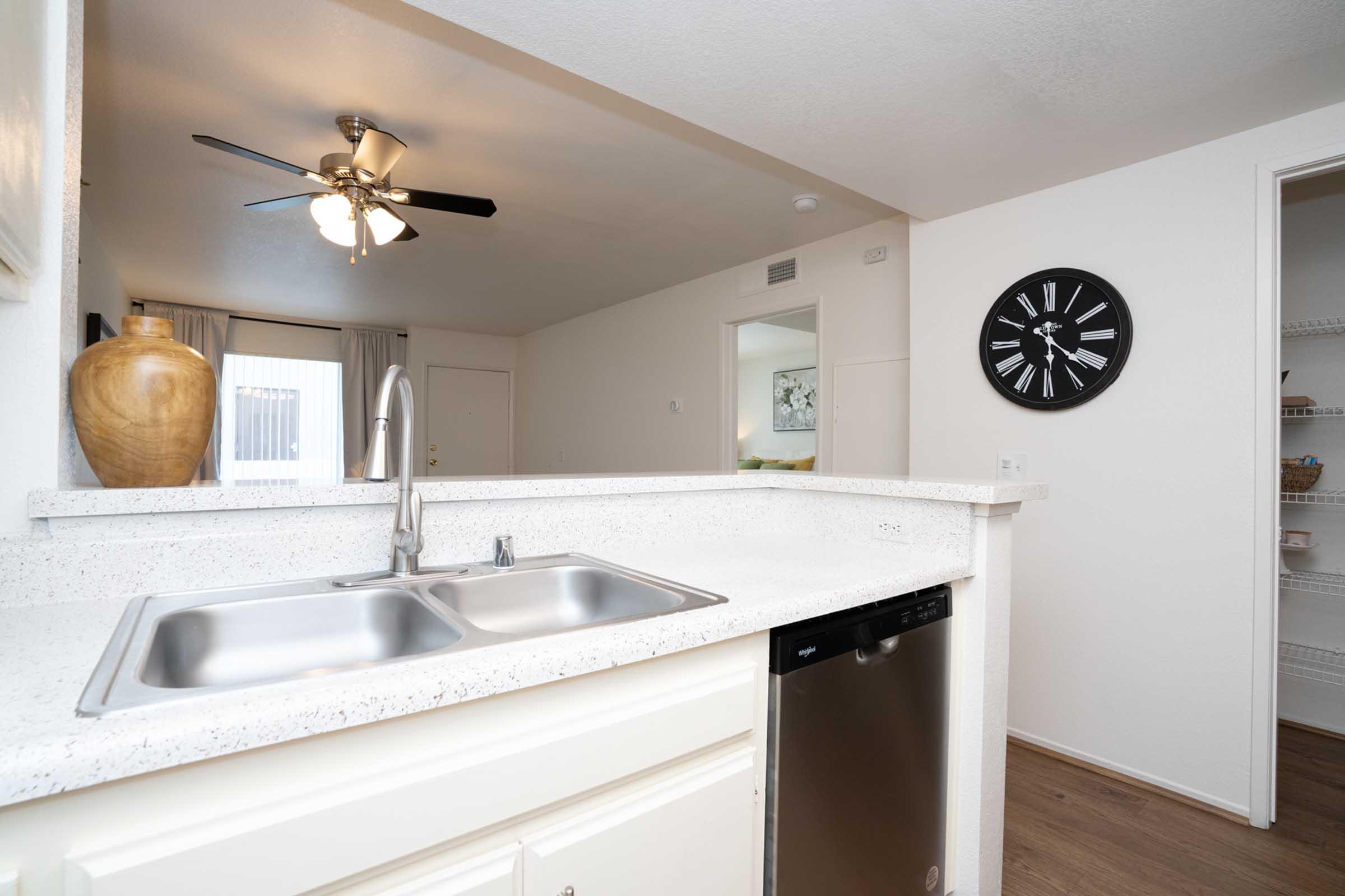 A modern kitchen with a white countertop and stainless steel sink, featuring a dishwasher and a ceiling fan. The background shows a living space with light-colored walls and a wall clock. Natural light filters through a nearby window, creating a bright and open atmosphere.