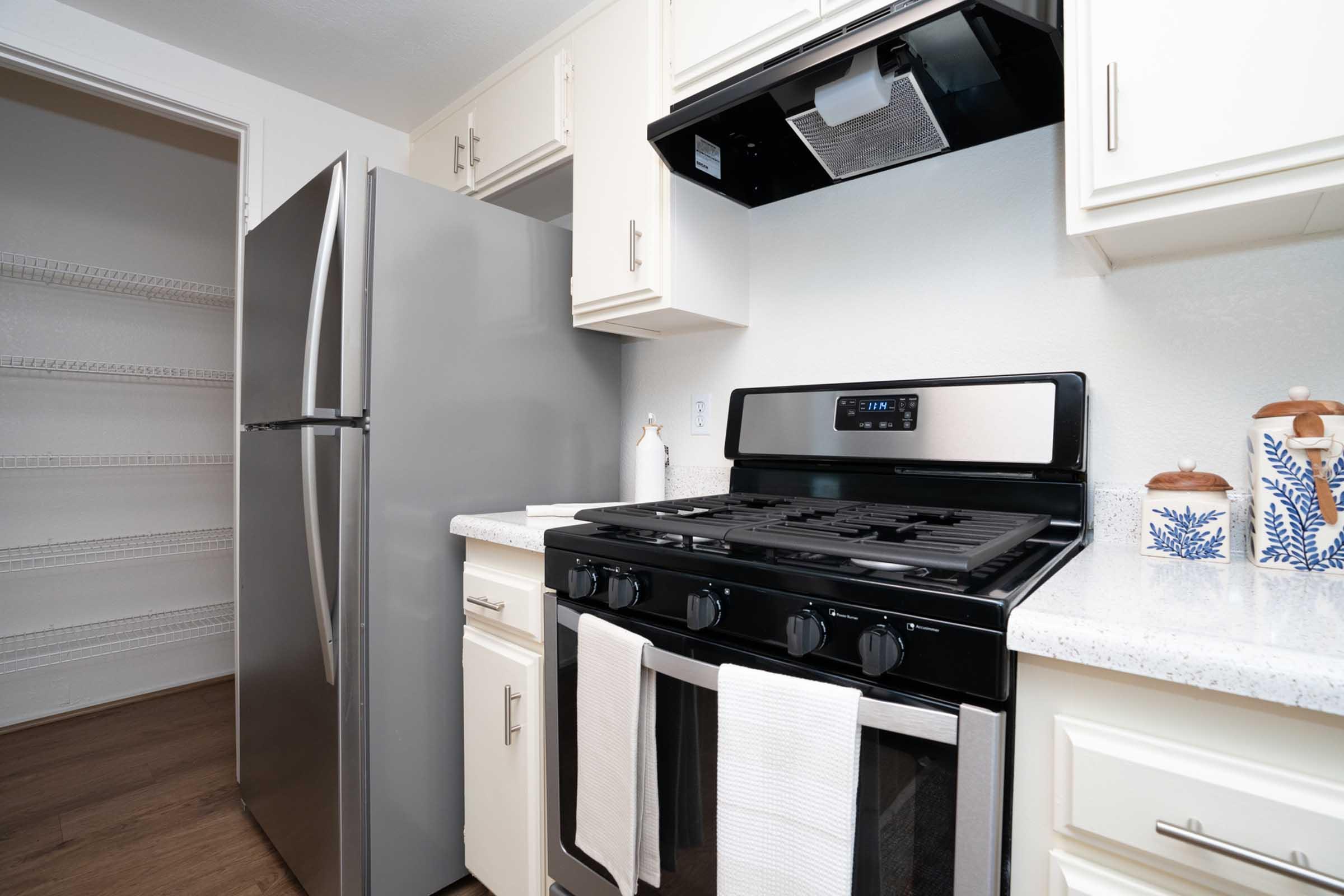 A modern kitchen featuring a stainless steel refrigerator, a gas stove with a black range hood, and white cabinets. The countertop is a light color, and there are two dish towels hanging in front of the stove. A pantry is visible in the background, with shelves ready for storage.