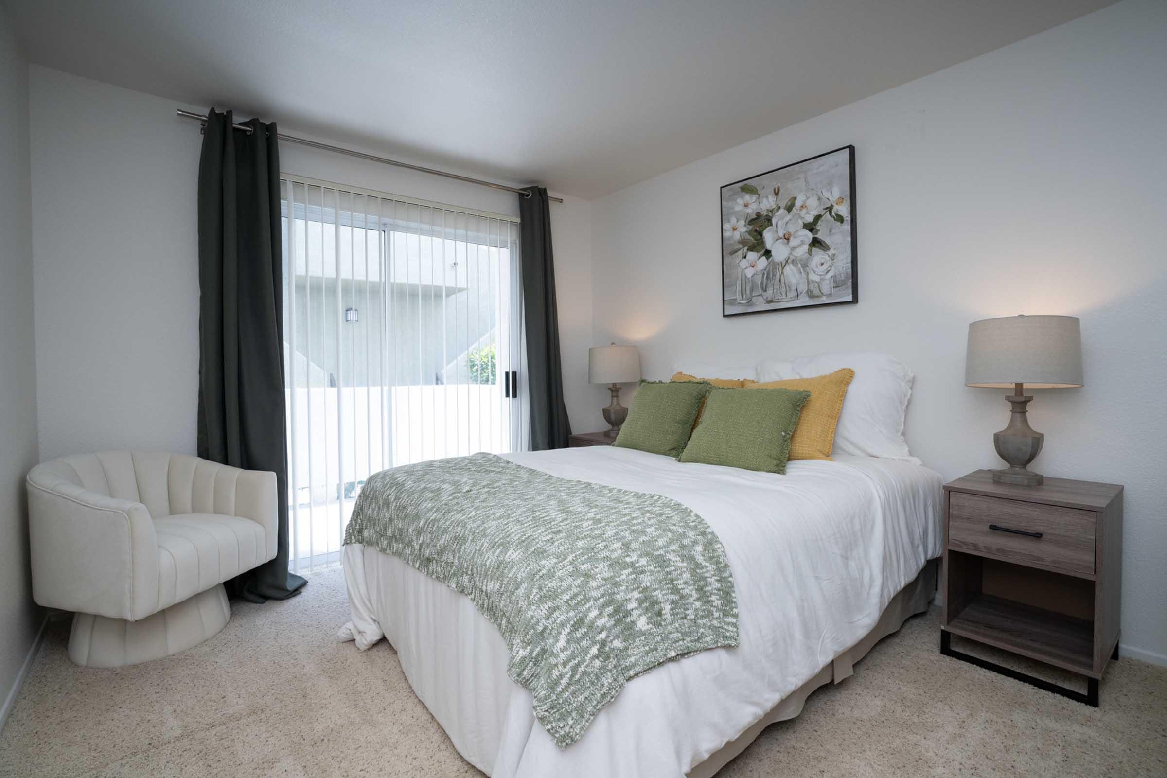 Cozy bedroom featuring a queen-sized bed with white bedding and green accent pillows. A modern armchair is positioned next to the bed. There are two bedside lamps and a floral artwork on the wall. Large sliding glass doors lead to an outdoor area, with sheer curtains allowing natural light.