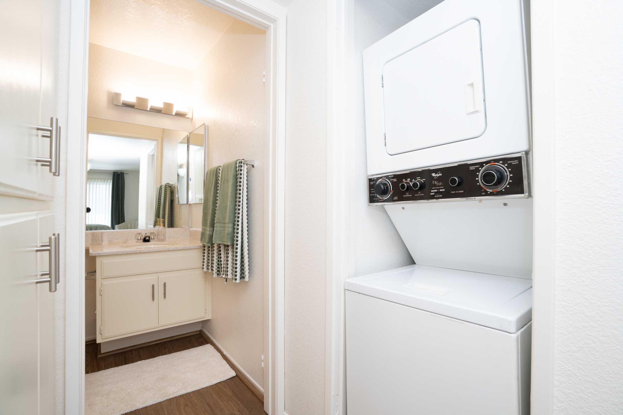 A view of a laundry area with a stacked washer and dryer. The space features a doorway leading to a well-lit bathroom, which includes a mirror, sink, and towel rack with green towels. The flooring is dark wood, and the walls are painted in light colors.