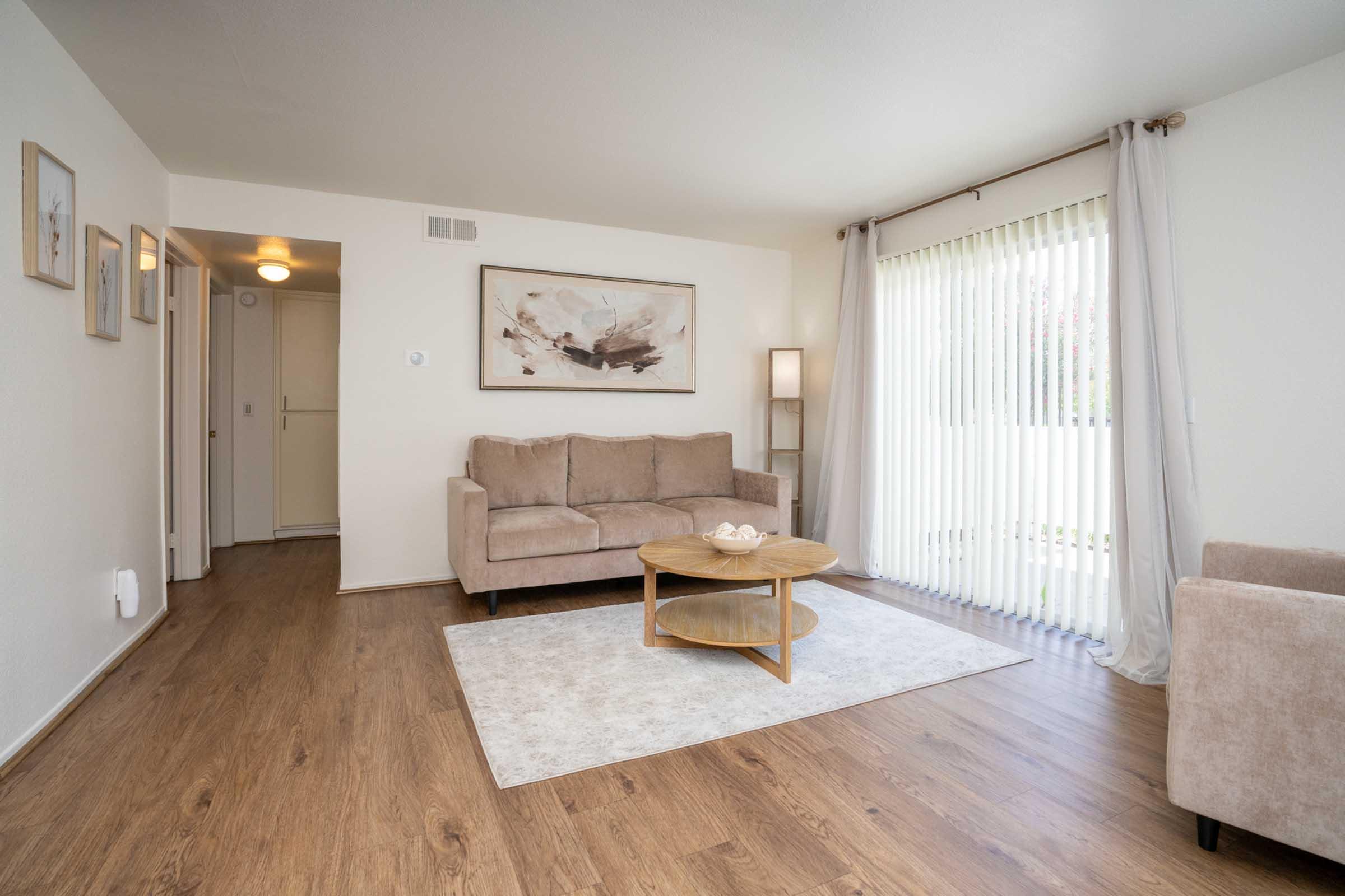 A cozy living room featuring a light brown sofa, a round coffee table with a decorative bowl, and a large window covered with vertical blinds. The room is adorned with framed artwork on the walls, soft lighting, and a neutral color palette, creating a warm and inviting atmosphere.